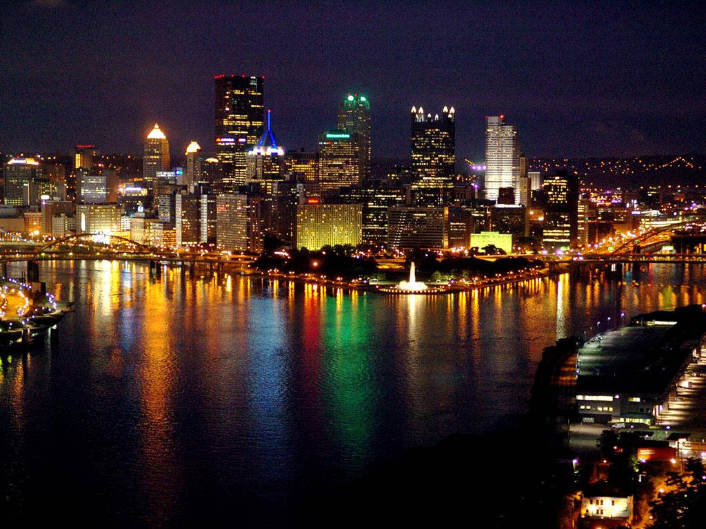 夜空 桌布 壁紙 — 一個耀眼的夜景城市景觀，摩天大樓在黑暗的天空中閃耀，它們的燈光倒映在下方平靜的河流中。