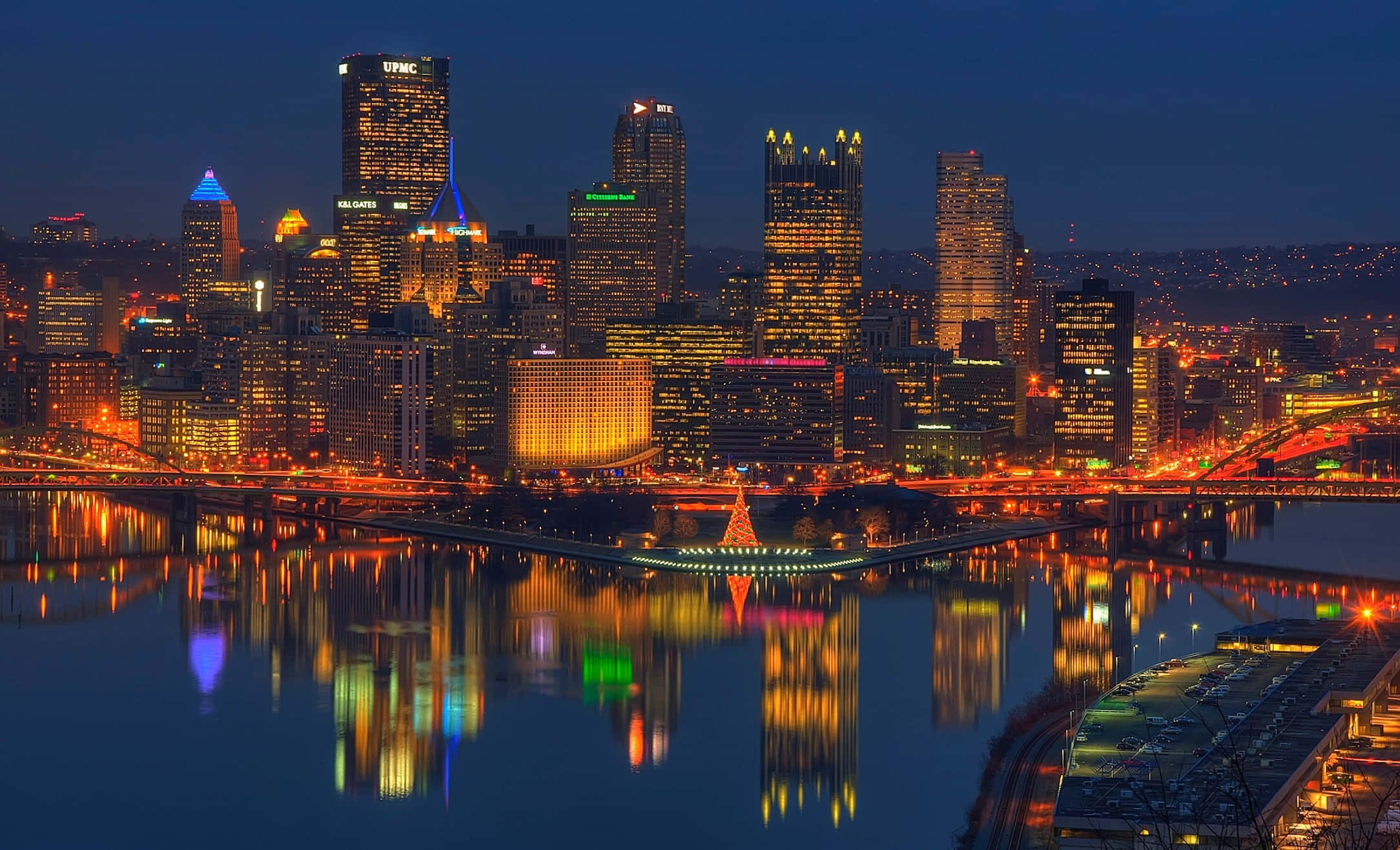 Pittsburgh Skyline 桌布 壁紙 — 一個耀眼的夜景城市景觀，摩天大樓在黑暗的天空中閃耀，它們的燈光完美地倒映在下方平靜的河流中。