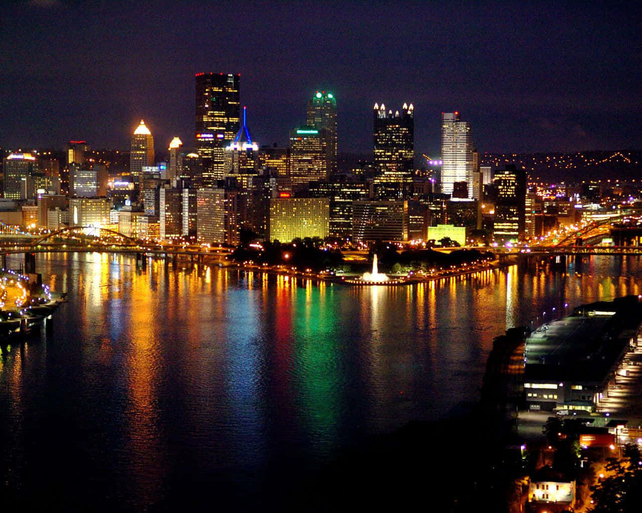 Pittsburgh Skyline 桌布 壁紙 — 一個耀眼的夜景城市景觀，摩天大樓在黑暗的天空中閃耀，它們的燈光生動地反射在下方平靜的河流上。