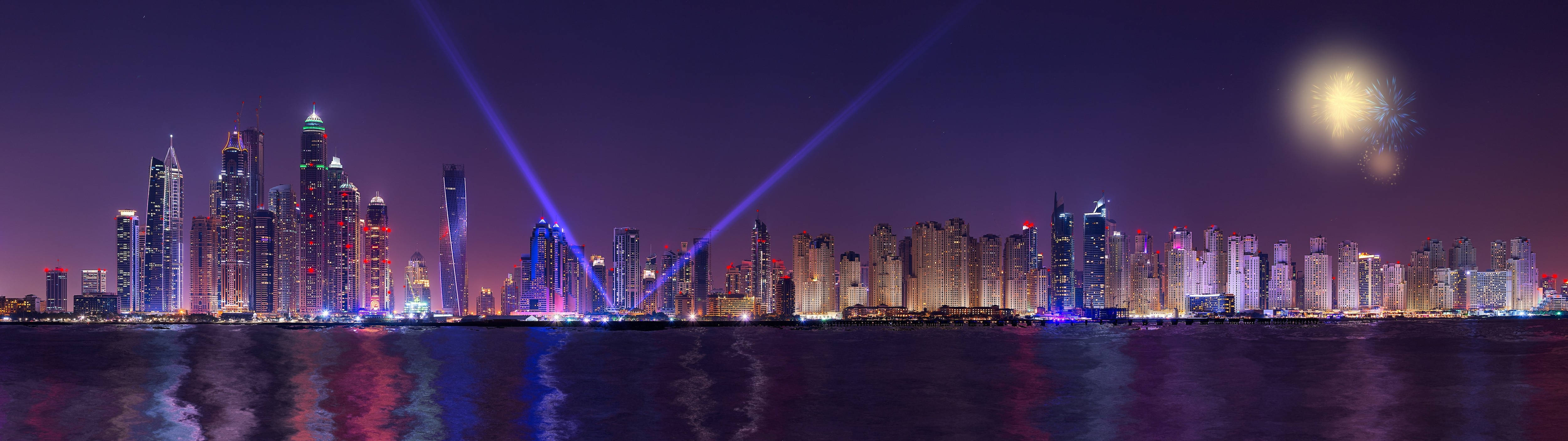 4K ultra widescreen 桌布 壁紙 — 一個耀眼的夜景城市景觀，高聳的摩天大樓被五彩繽紛的燈光和雷射光束照亮，在水面上反射，頭頂上綻放煙火。