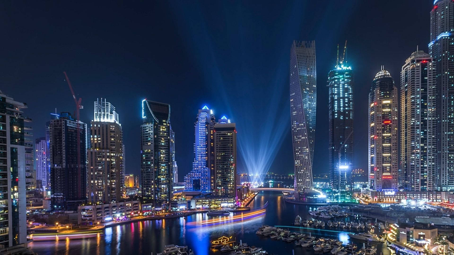 Dubai 桌布 壁紙 — 一個耀眼的夜景城市景觀，高聳的摩天大樓被鮮豔的燈光和光束照亮，倒映在滿是船隻的水道上。