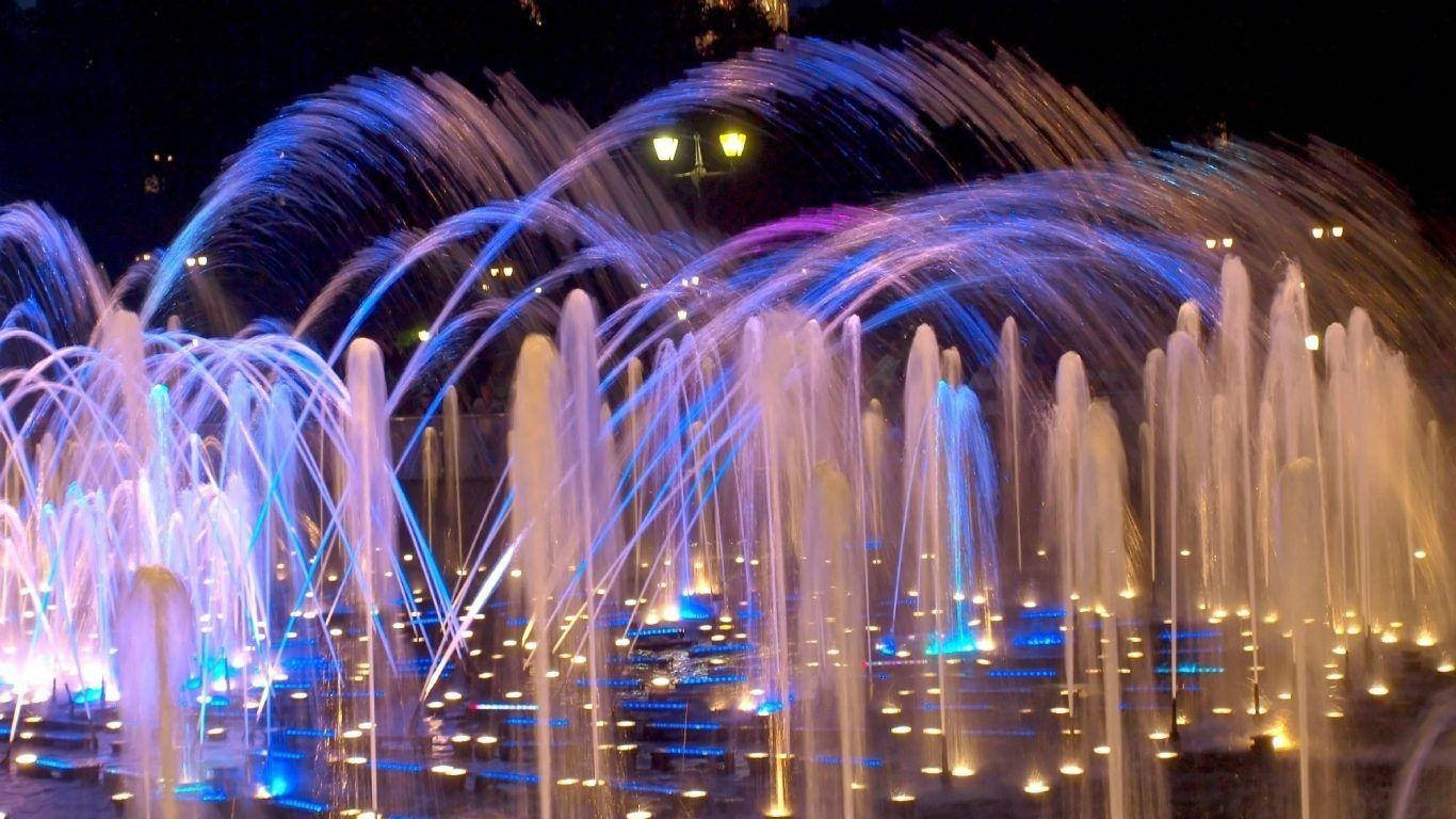 水 桌布 壁紙 — 一個耀眼的夜間噴泉表演，弧形水柱在鮮豔的藍色和紫色燈光下閃爍。