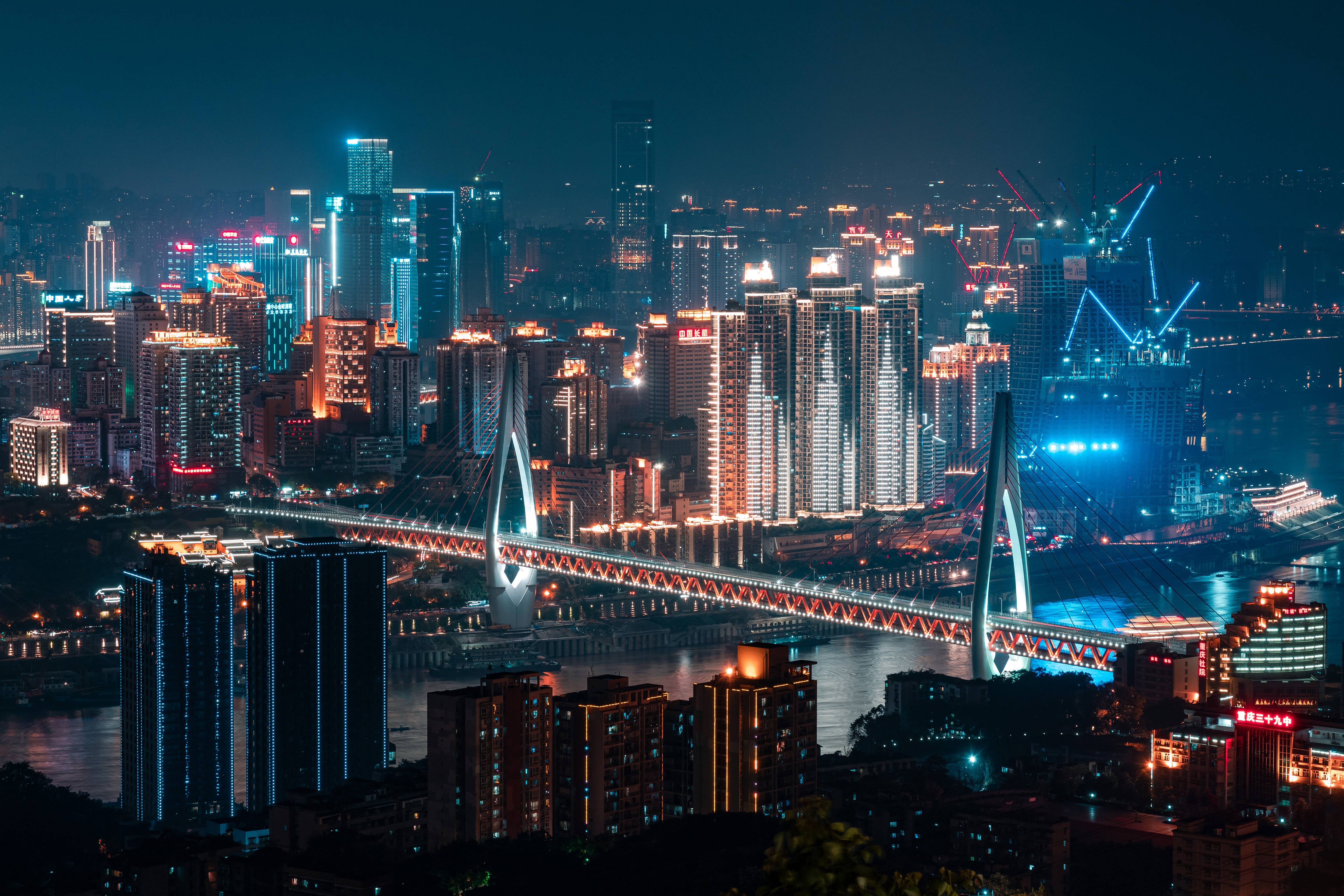 Chongqing 桌布 壁紙 — 一個耀眼的夜間城市景觀，有一座發光的橋樑橫跨河流，周圍是閃爍著鮮豔霓虹色彩的摩天大樓。