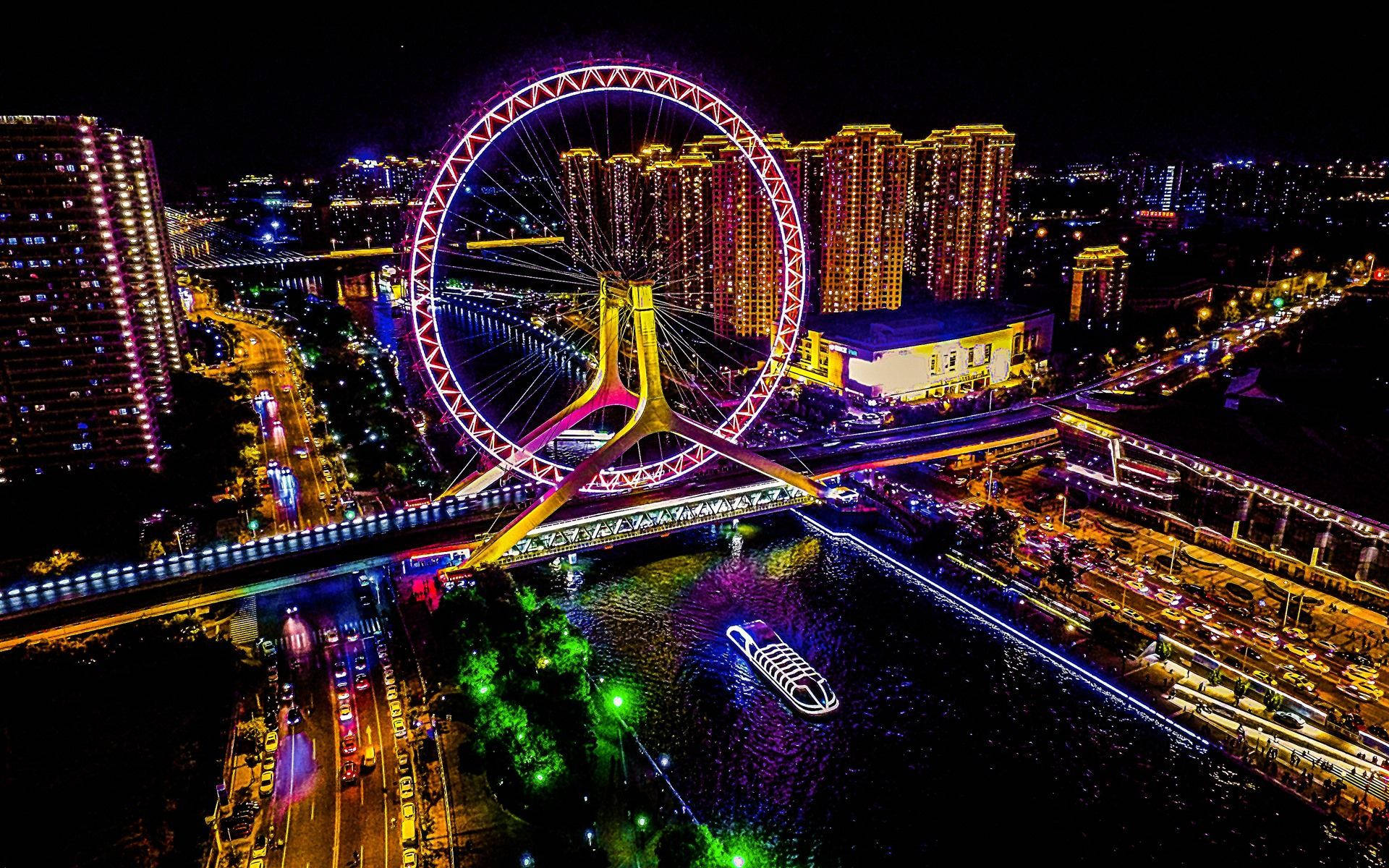 Tianjin 桌布 壁紙 — 一個耀眼的夜間城市景觀，特色是橫跨河流的明亮摩天輪，下方有發光的建築物和一艘亮燈的船。