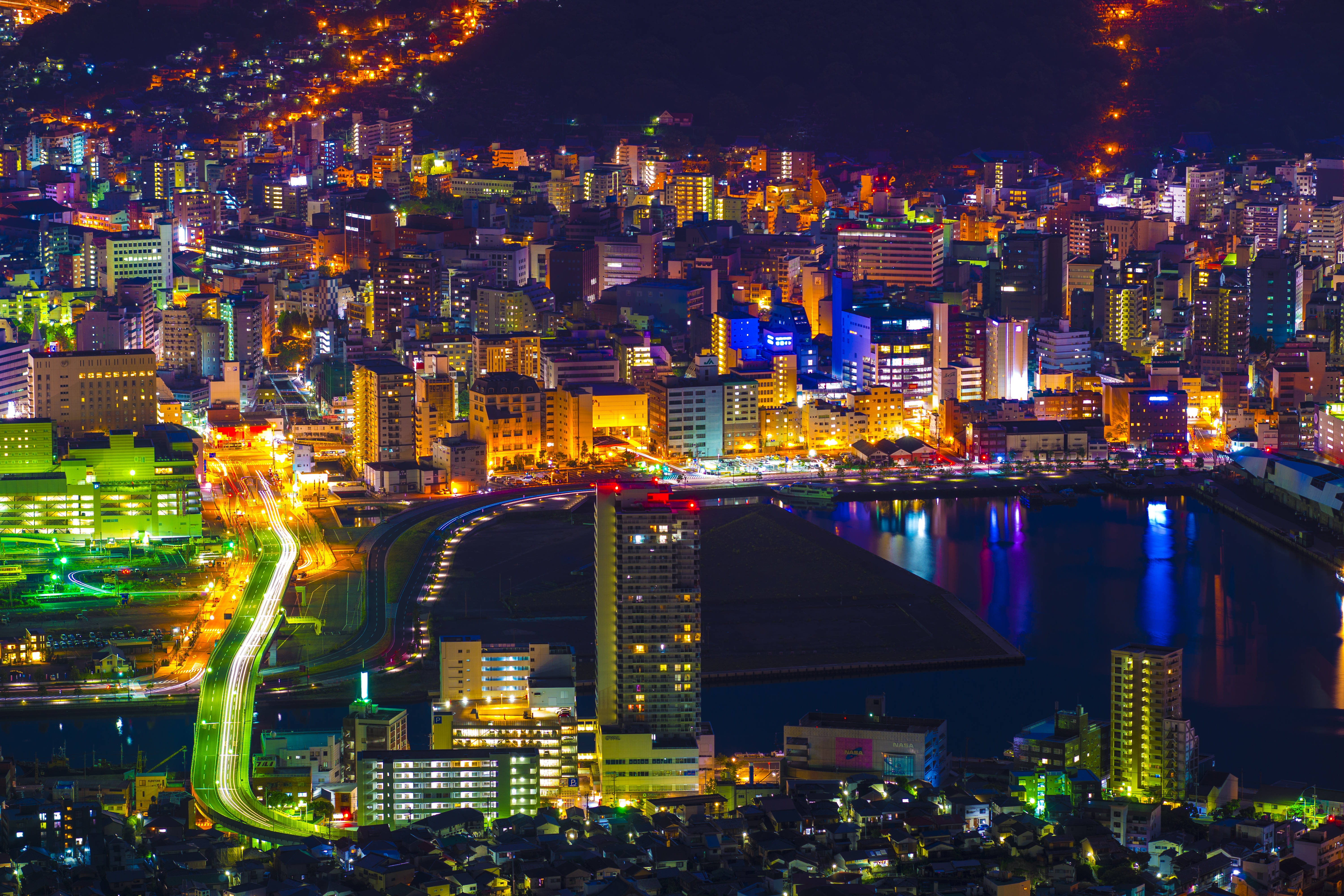 日本 桌布 壁紙 — 一個耀眼的夜間城市景觀，閃爍的霓虹燈光倒映在水面上，車流的光跡照亮蜿蜒的道路。