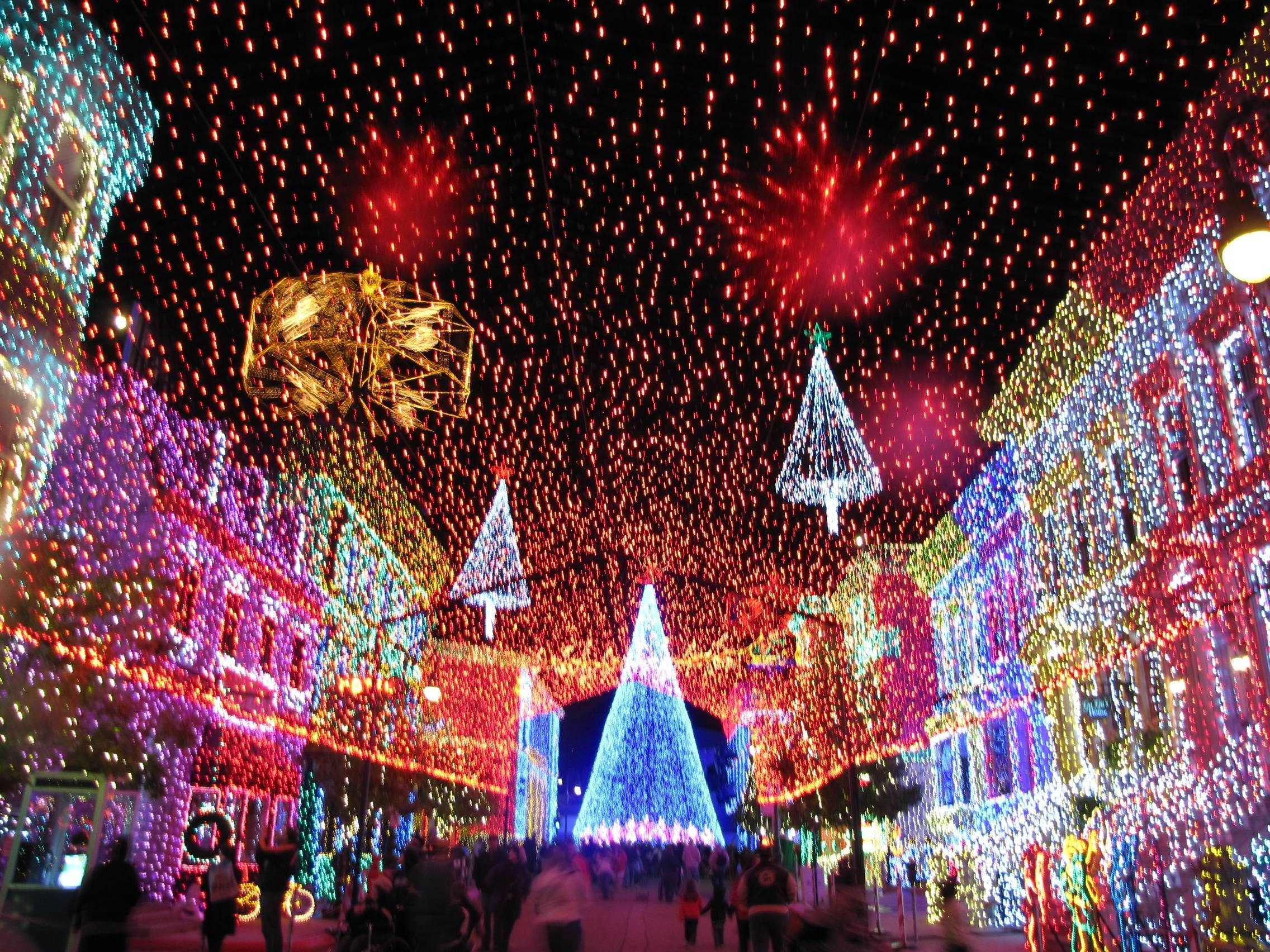 Festive 桌布 壁紙 — 一個耀眼的夜間街頭節日，建築物和樹木被五彩繽紛的燈光覆蓋，頭頂上煙花綻放。