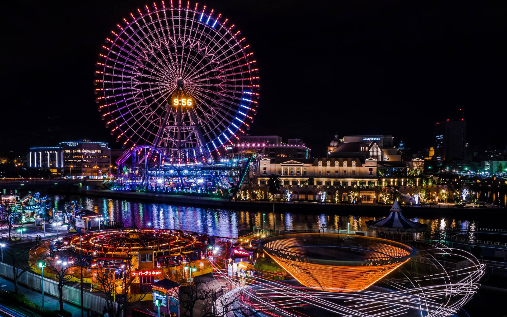Yokohama 桌布 壁紙 — 一個耀眼的夜間遊樂園，巨大的燈光摩天輪倒映在水中，周圍是閃爍的遊樂設施和建築物。