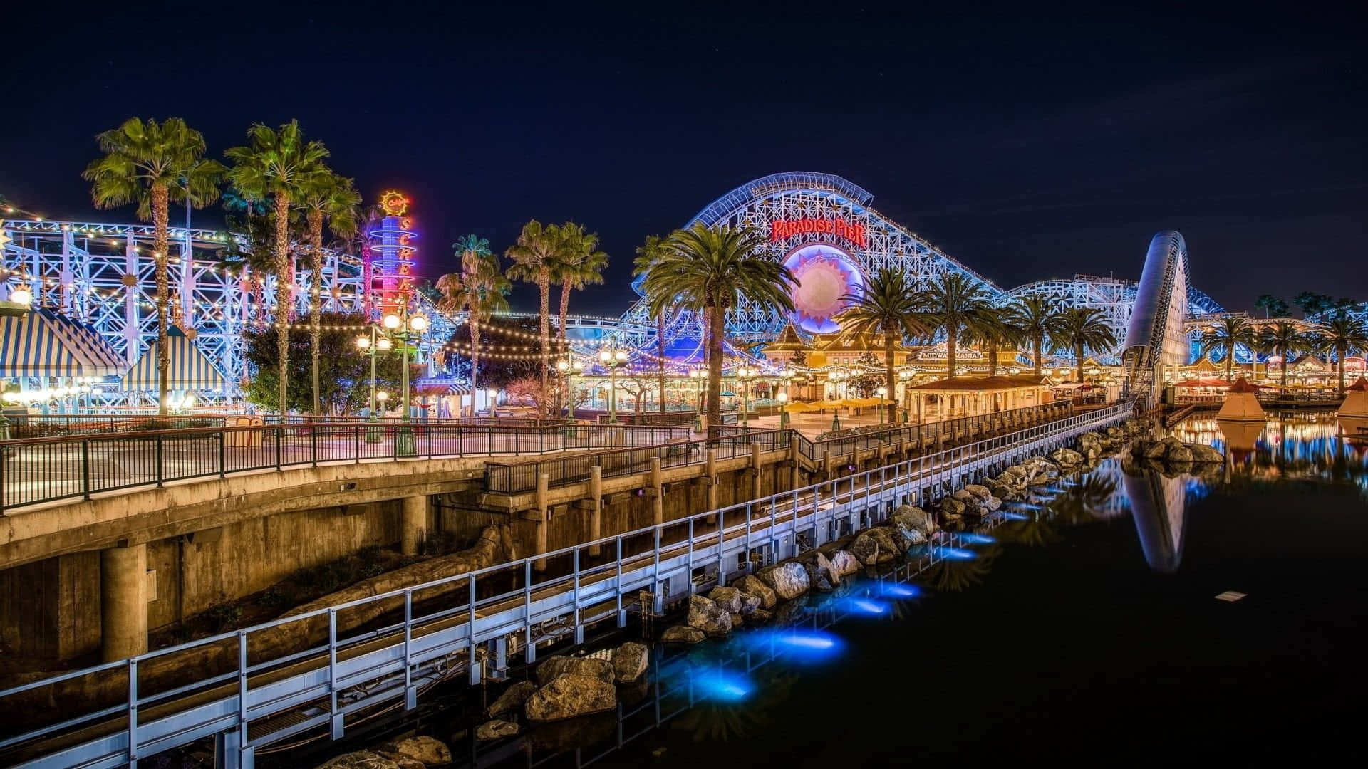 Roller Coaster 桌布 壁紙 — 一個耀眼的夜間遊樂園，有發光的雲霄飛車、棕櫚樹和倒映在平靜水面上的鮮豔燈光。