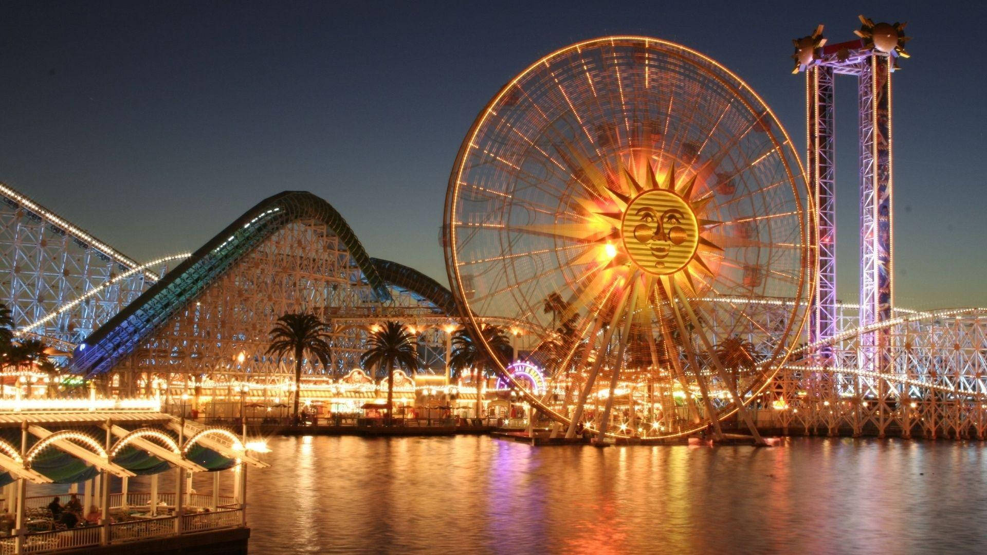 Theme Park 桌布 壁紙 — 一個耀眼的夜間遊樂園，發光的太陽主題摩天輪和雲霄飛車倒映在平靜的水面上。