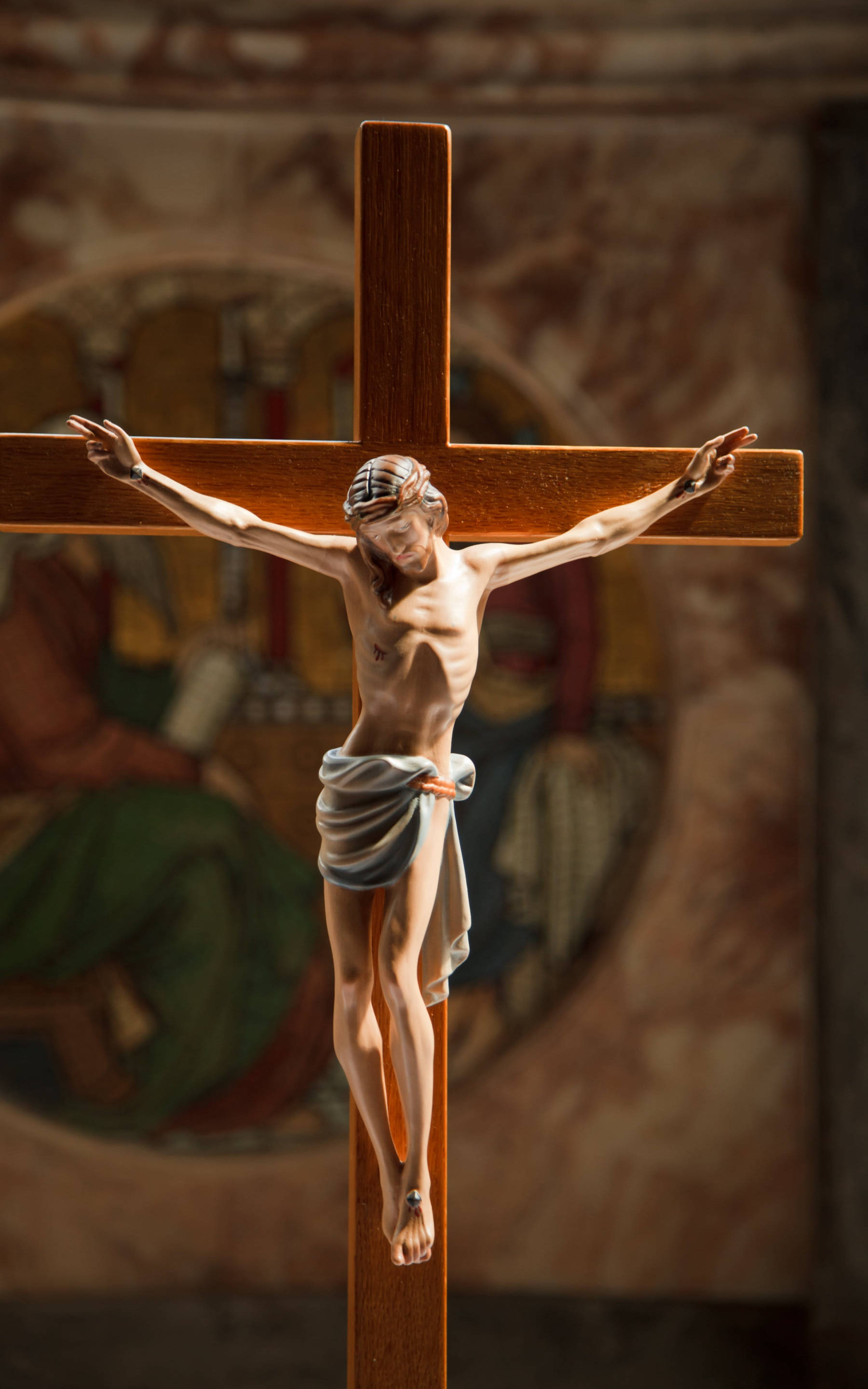 Jesus Christ 桌布 壁紙 — 一個耶穌基督的木製十字架雕像掛在燈光柔和的教堂裡，背景有一幅褪色的宗教壁畫。