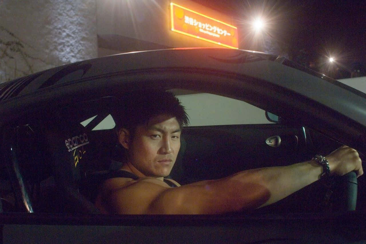Fast And Furious 桌布 壁紙 — 一個肌肉男在夜晚霓虹燈下緊握一輛時尚黑色跑車的方向盤。