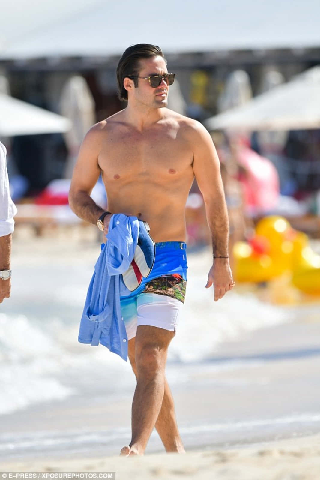 Spencer Matthews 桌布 壁紙 — 一個肌肉男戴著太陽眼鏡，赤腳在陽光沙灘上行走，拿著一件藍色襯衫和人字拖。