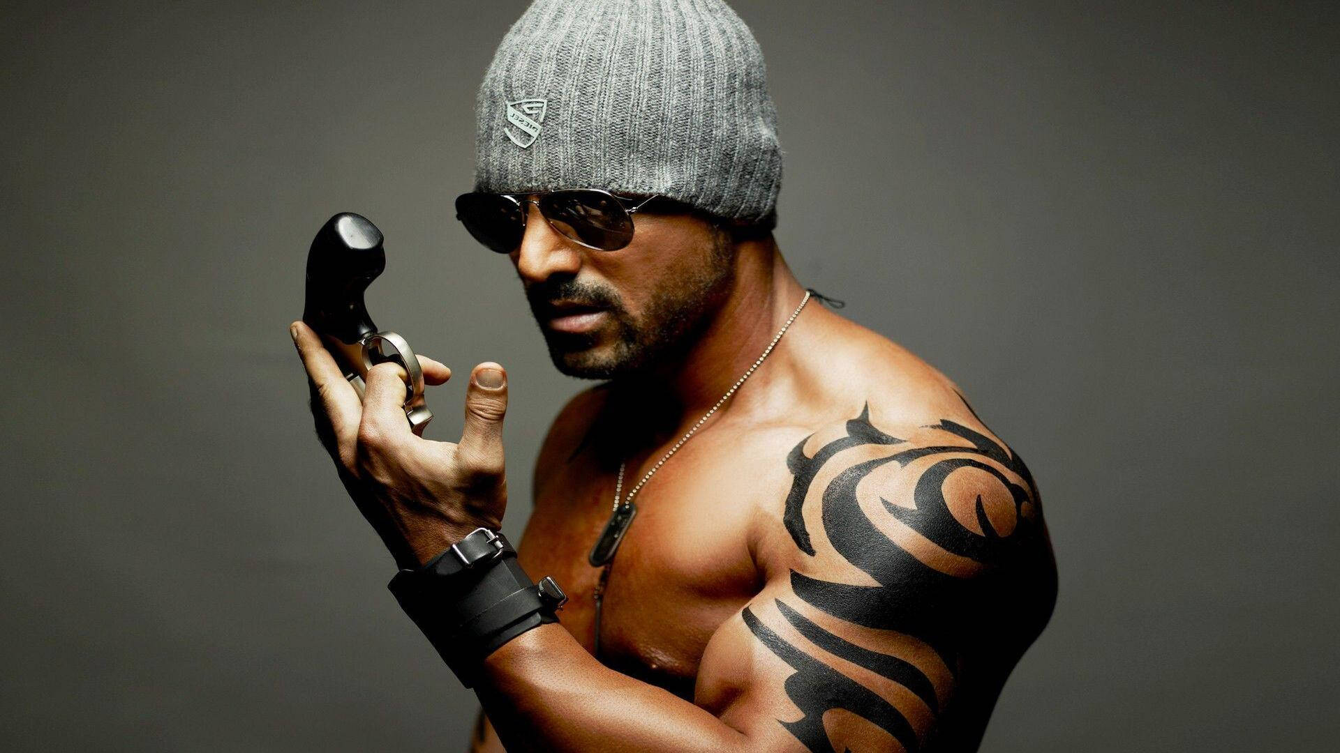 John Abraham 桌布 壁紙 — 一個肌肉發達、帶有部落紋身的男人，戴著毛帽和太陽眼鏡，在灰色背景前拿著左輪手槍。