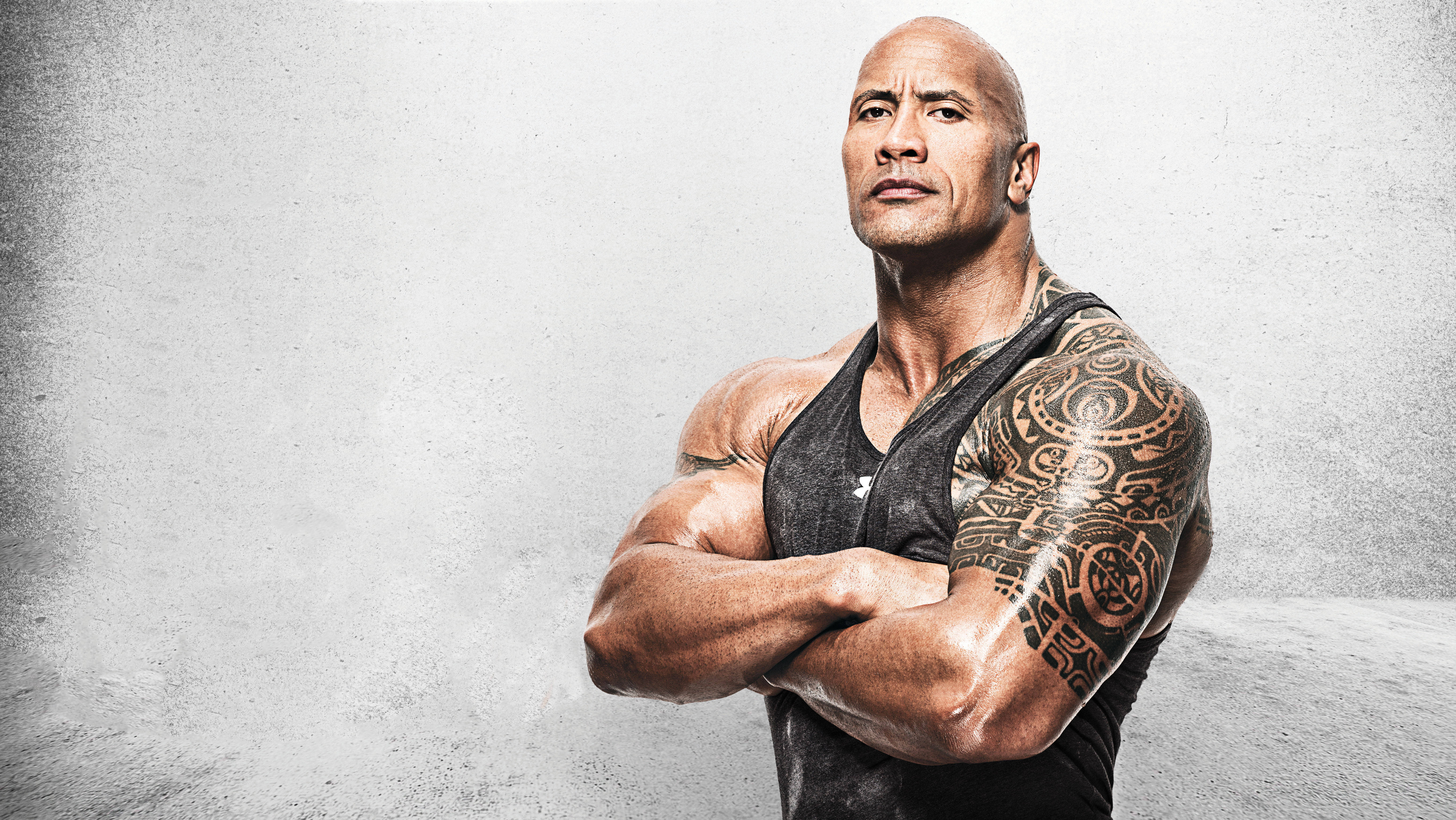 Dwayne 桌布 壁紙 — 一個肌肉發達、帶有部落紋身的男人自信地交叉雙臂，靠著紋理灰色牆壁站立。