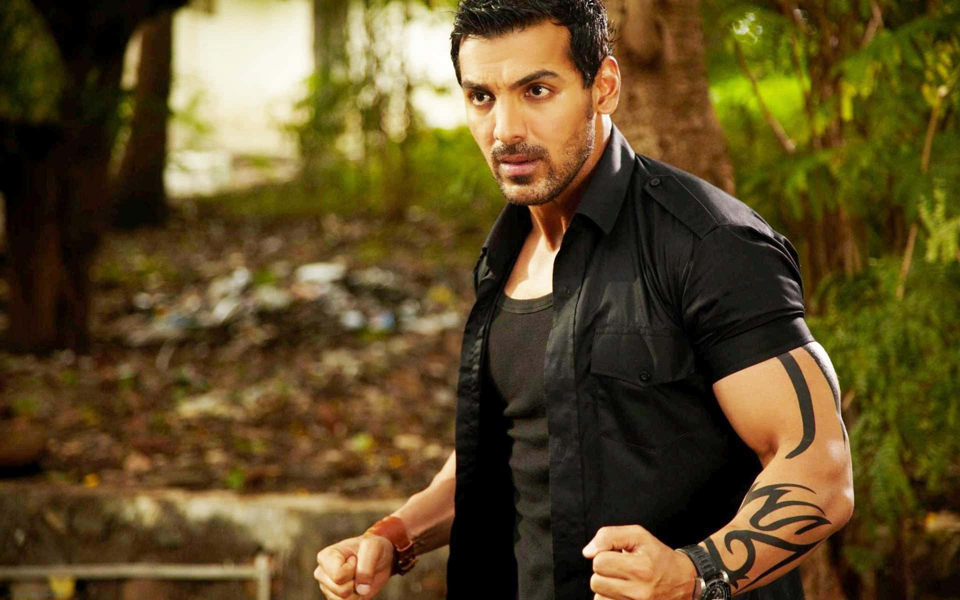 John Abraham 桌布 壁紙 — 一個肌肉發達、手臂有刺青的男人站在陽光明媚的森林中，握緊拳頭，身穿黑色襯衫。