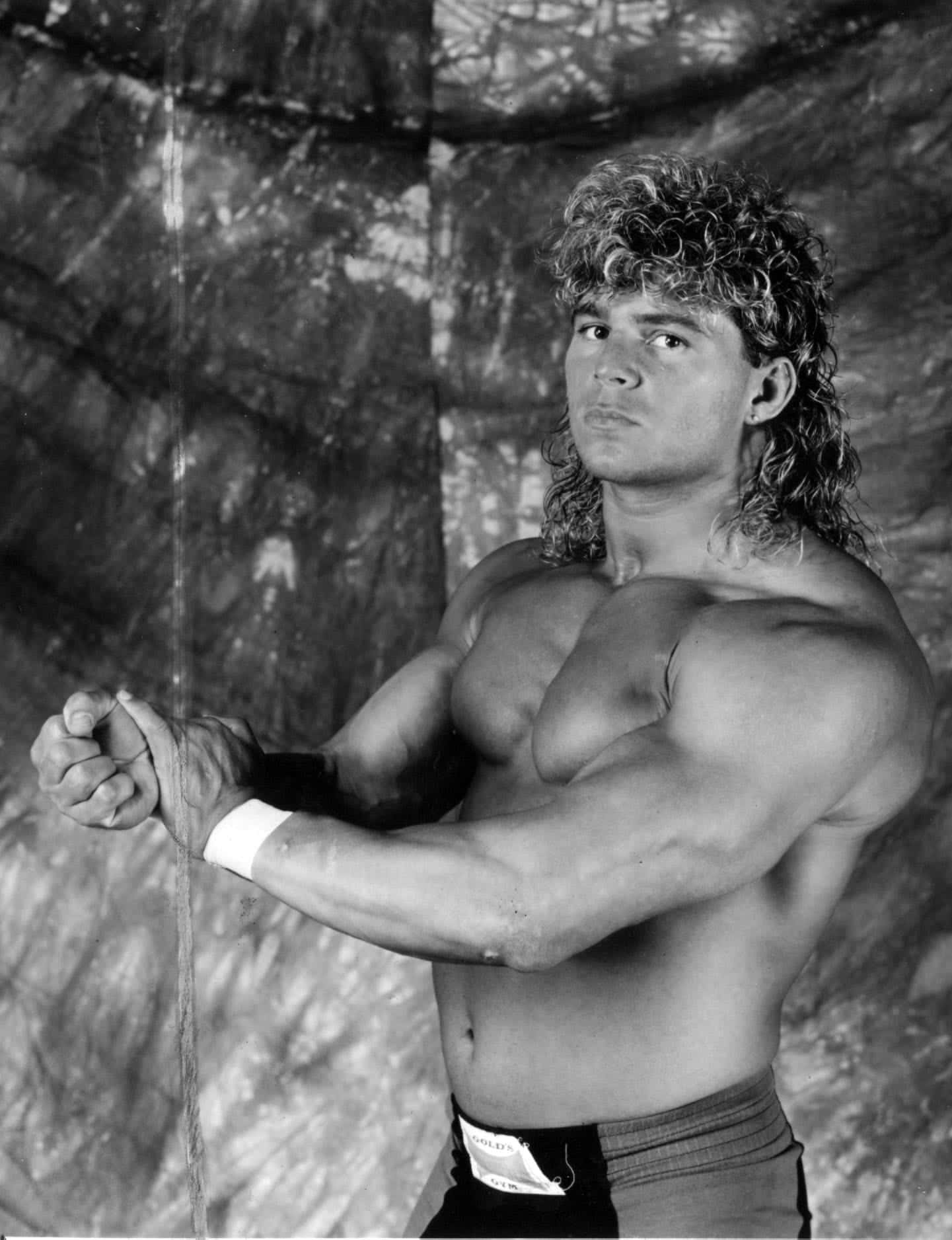 WWE 桌布 壁紙 — 一個肌肉發達、擁有濃密捲髮的男人，穿著摔角褲和護腕，在紋理背景前彎曲手臂。