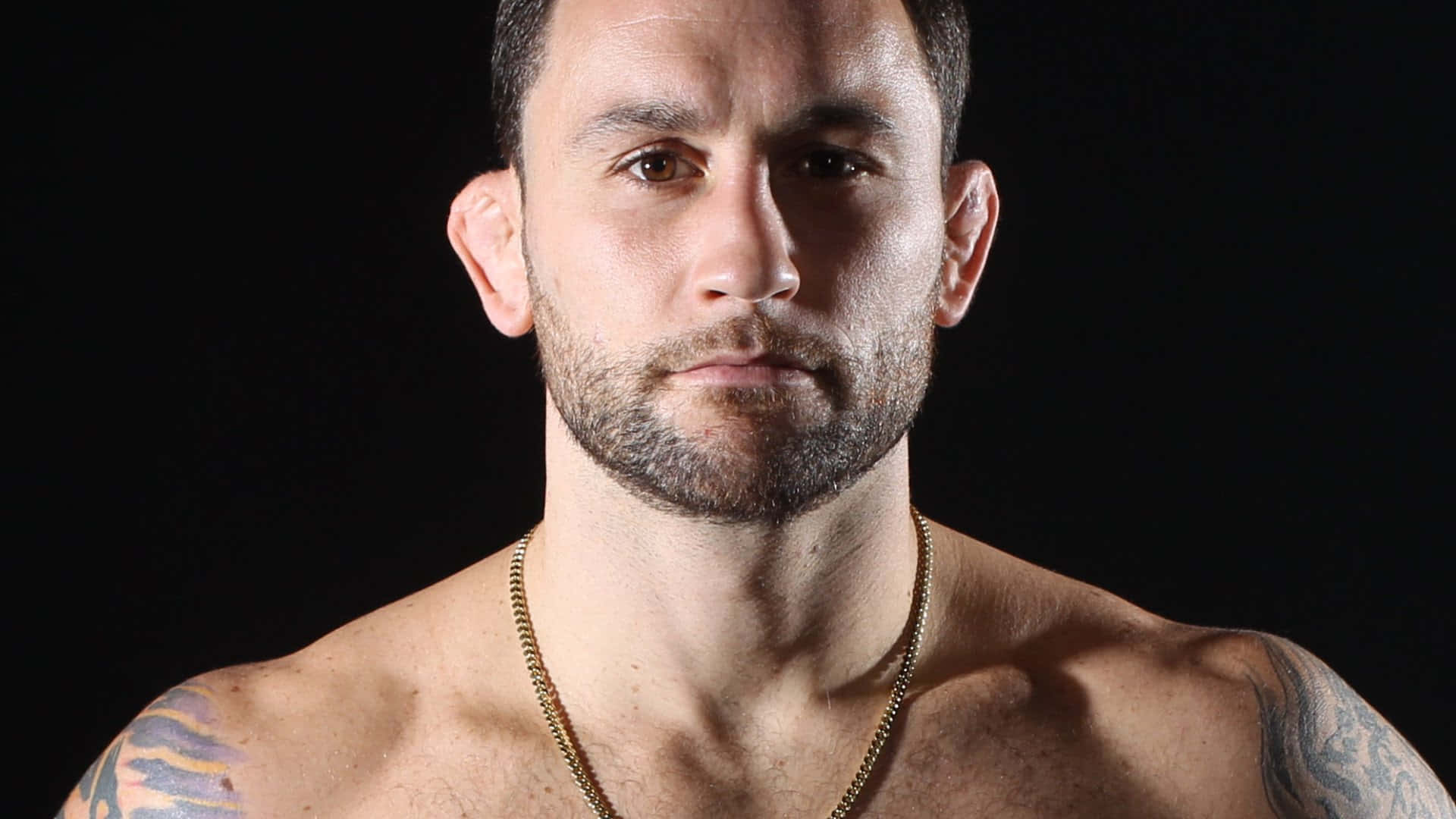 UFC 桌布 壁紙 — 一個肌肉發達、有刺青和戴金項鍊的男人，在黑色背景前強烈地凝視。