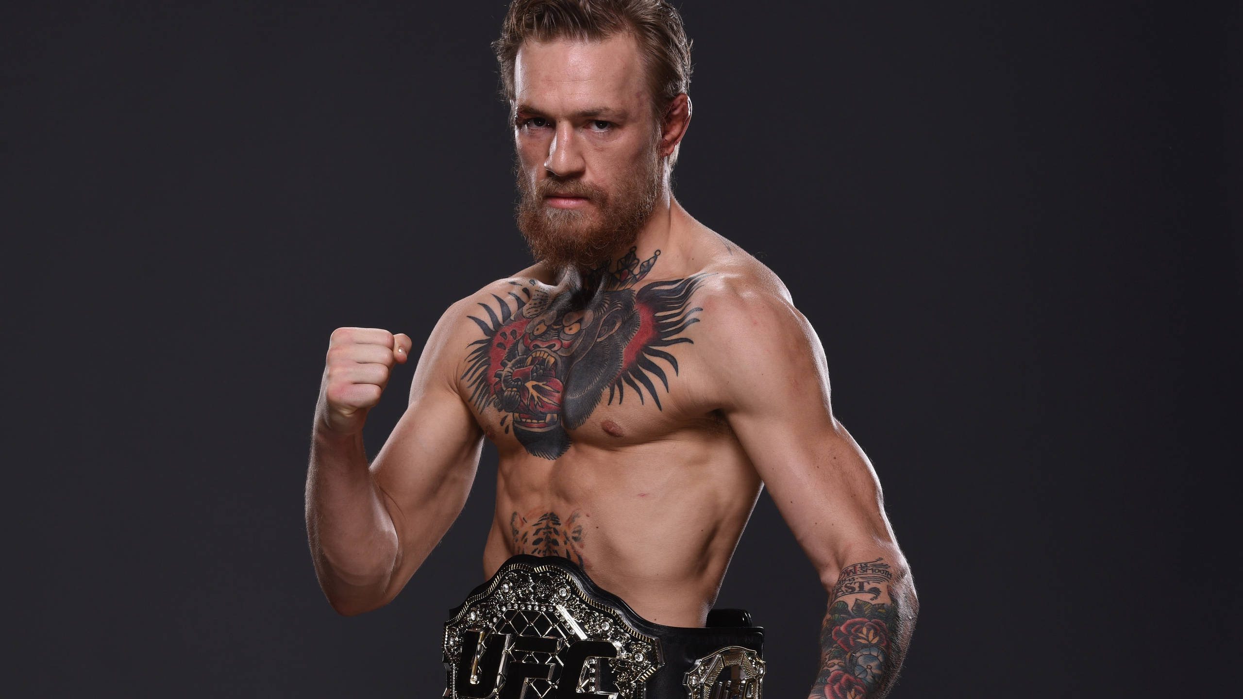Mcgregor 桌布 壁紙 — 一個肌肉發達、有鬍鬚的戰士，胸口有紋身，在黑暗的攝影棚背景下握緊拳頭，手持UFC冠軍腰帶。