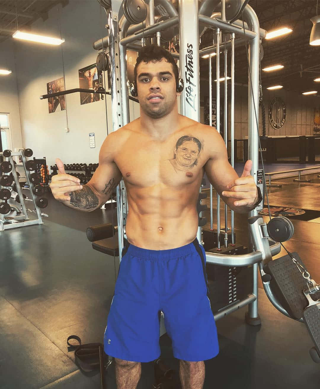 UFC 桌布 壁紙 — 一個肌肉發達、沒穿衣服的男人，身上有刺青，在燈光充足的健身房裡，周圍是舉重機，他舉起兩個大拇指。