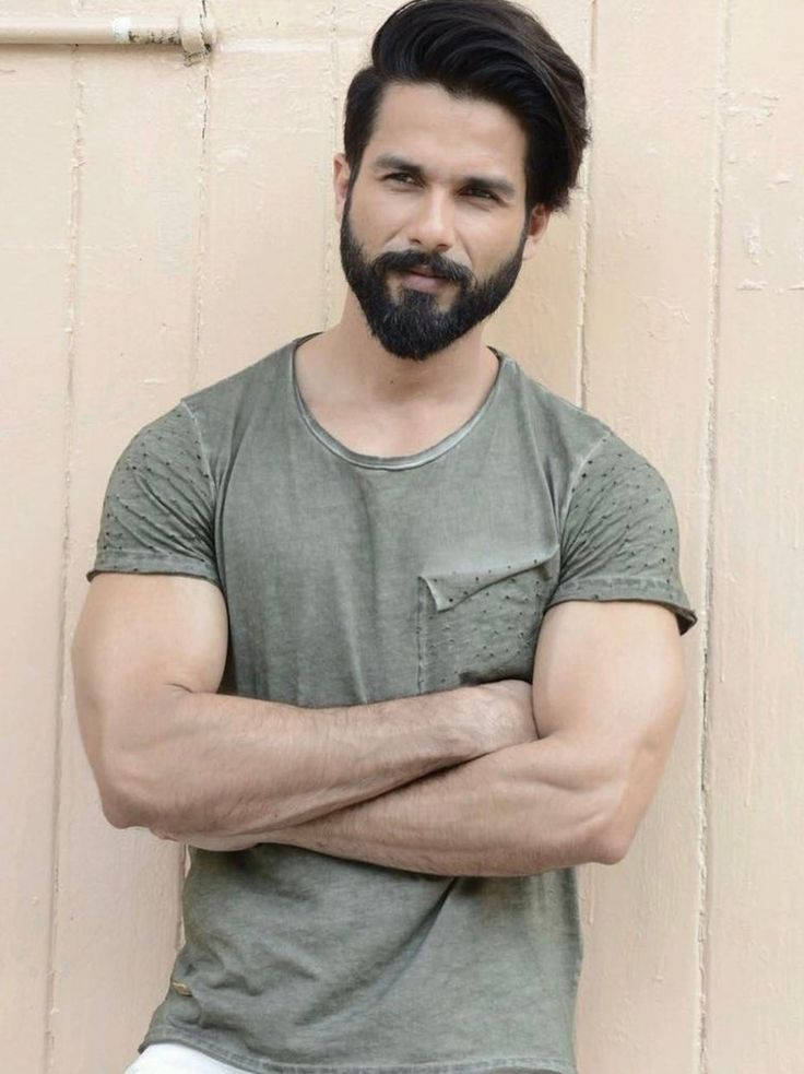 Shahid Kapoor 桌布 壁紙 — 一個肌肉發達、留著濃密鬍鬚、頭髮造型時尚的男人自信地交叉雙臂，靠著米色牆壁站立，身穿褪色綠色T恤。