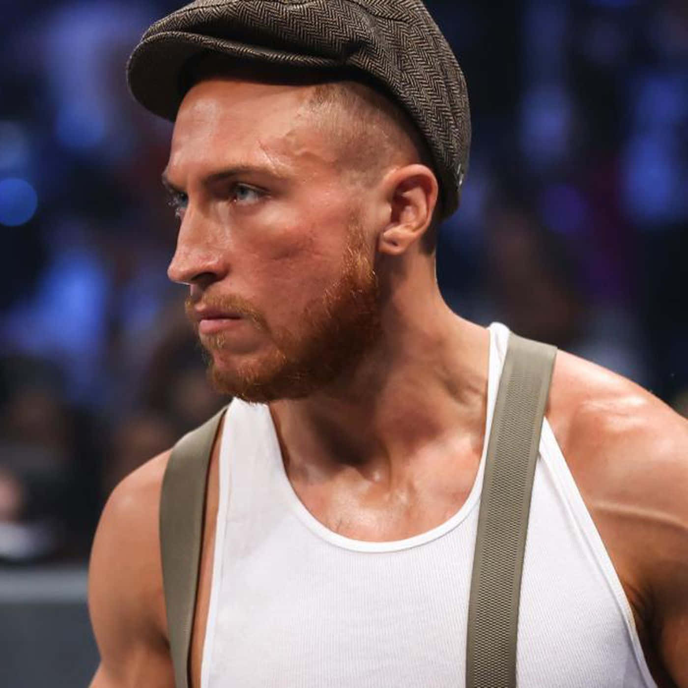 WWE 桌布 壁紙 — 一個肌肉發達、留著紅鬍子、戴著平頂帽的男人在明亮的燈光下在摔角場上凝視著。