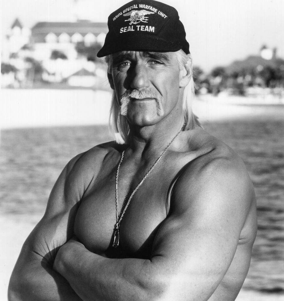 Hulk Hogan 桌布 壁紙 — 一個肌肉發達、留著金色長髮和鬍鬚的男人，赤裸上身站在沙灘上，戴著SEAL TEAM帽子和項鍊。