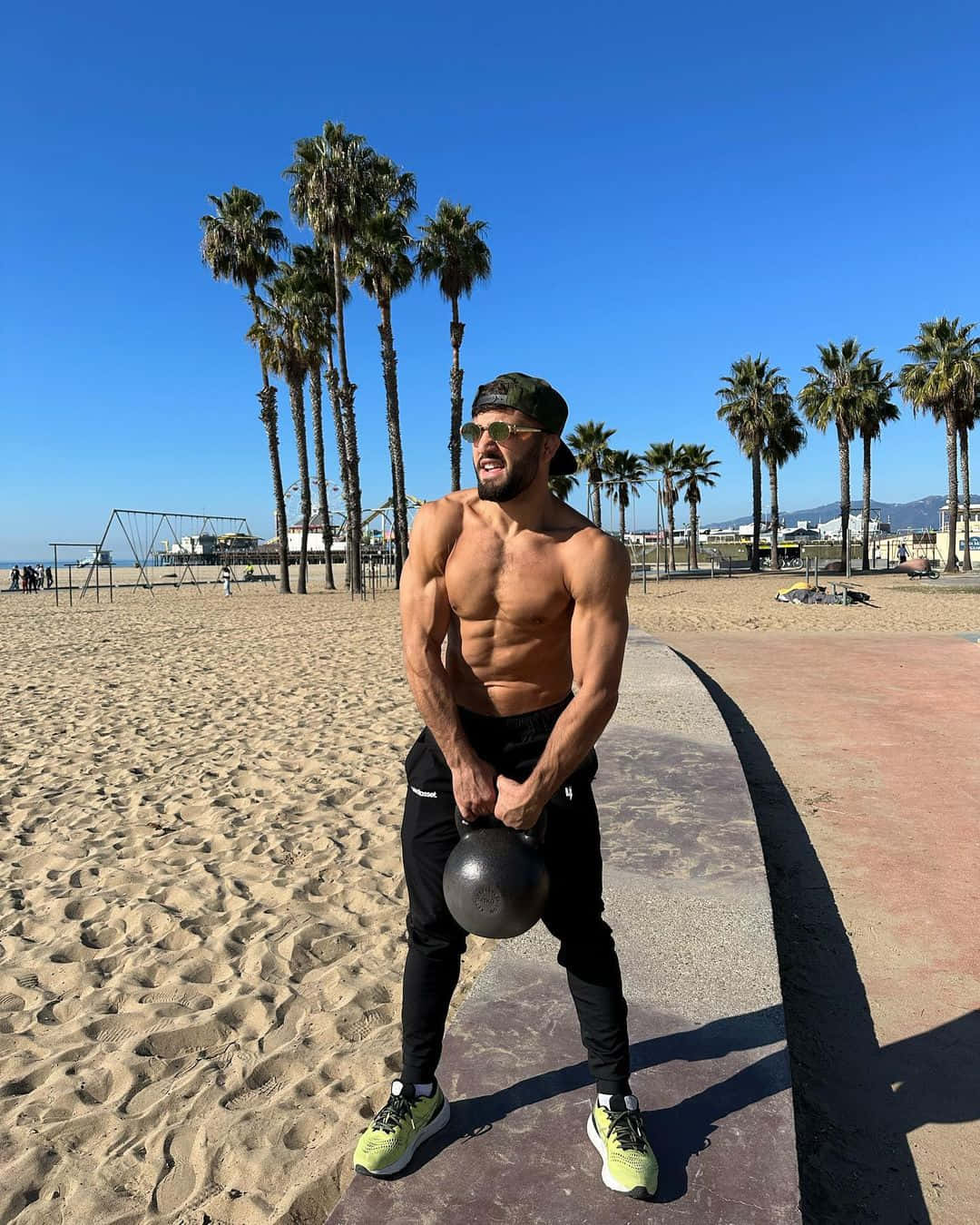 Fitness 桌布 壁紙 — 一個肌肉發達、留鬍子、戴帽子的男人在陽光明媚、棕櫚樹成蔭的海灘小徑上舉起壺鈴。