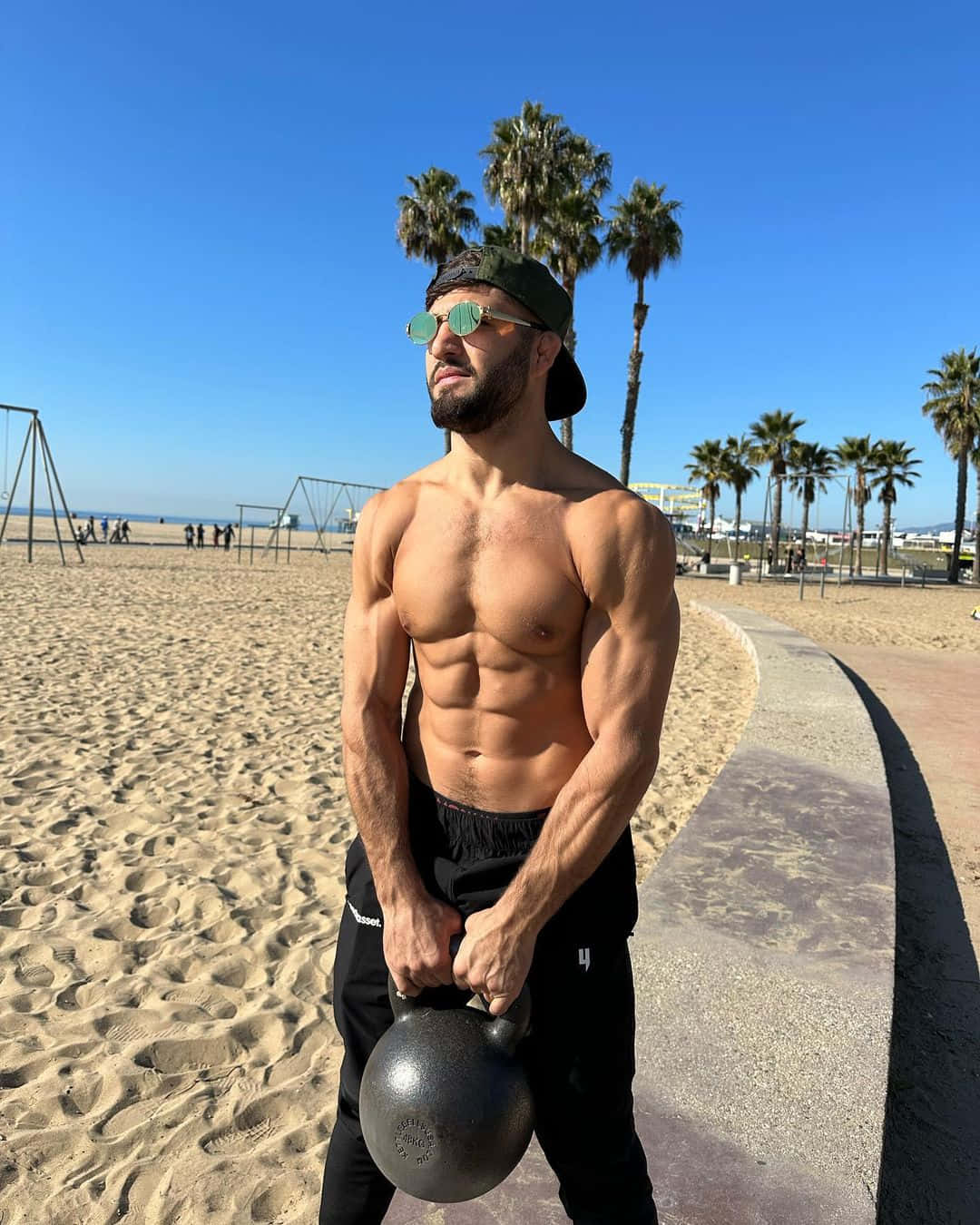 Fitness 桌布 壁紙 — 一個肌肉發達、留鬍子、戴綠色太陽眼鏡的男人，在陽光明媚、兩旁種滿棕櫚樹的海灘小徑上拿著壺鈴。