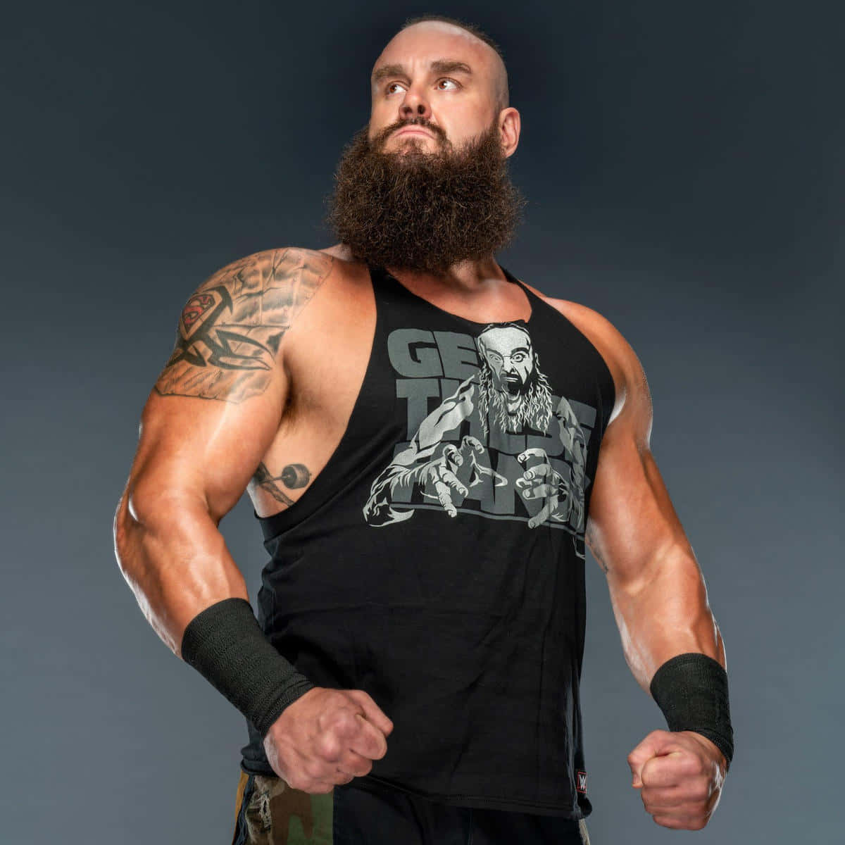 WWE 桌布 壁紙 — 一個肌肉發達、留鬍子的摔角手，身穿帶有圖案印花的黑色背心，在黑暗的攝影棚背景下握緊拳頭站立。