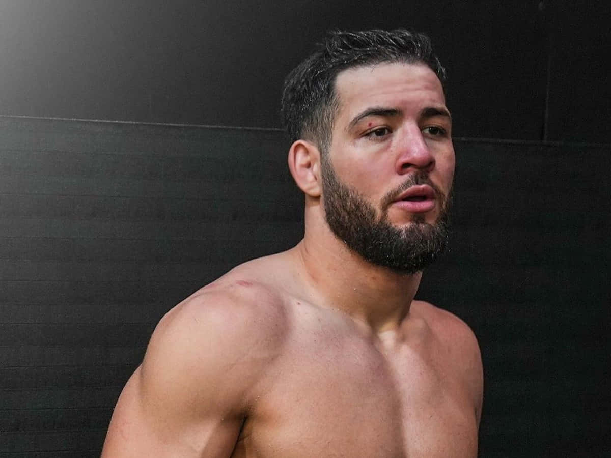 UFC 桌布 壁紙 — 一個肌肉發達、留鬍鬚的男人，神情專注，赤裸上身靠著深色紋理牆壁。