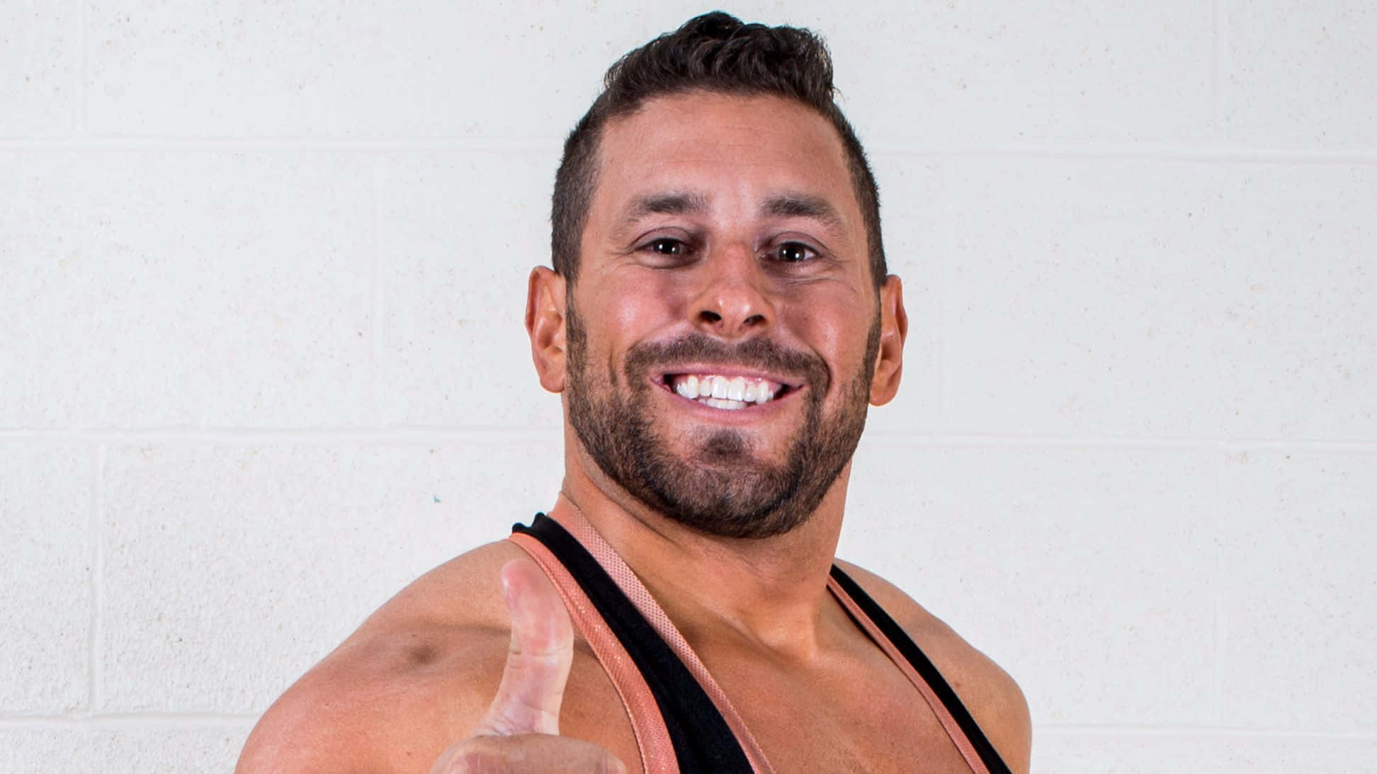 WWE 桌布 壁紙 — 一個肌肉發達、留鬍鬚的男人對著純白色磚牆咧嘴大笑，豎起大拇指。
