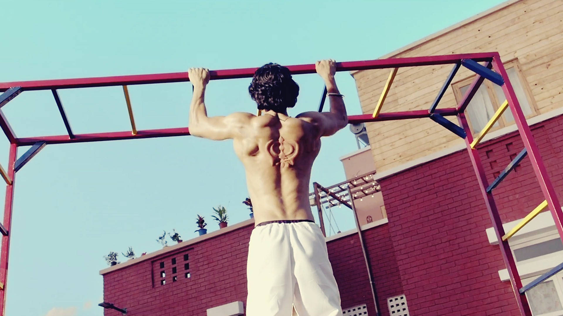 Fitness 桌布 壁紙 — 一個肌肉發達、背部有刺青的男人，在晴朗藍天下，以紅磚建築為背景，在色彩繽紛的戶外槓上做引體向上。