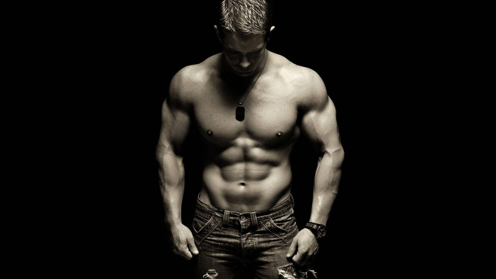 Fitness 桌布 壁紙 — 一個肌肉發達、腹肌分明的男人，赤裸上身，穿著牛仔褲和狗牌項鍊，站在黑色背景前。