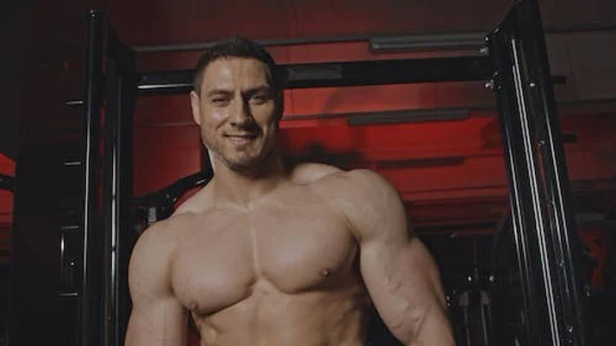 Exercise 桌布 壁紙 — 一個肌肉發達、自信微笑的男人，在健身房中赤裸上身，被紅色燈光照射在黑色器材上。