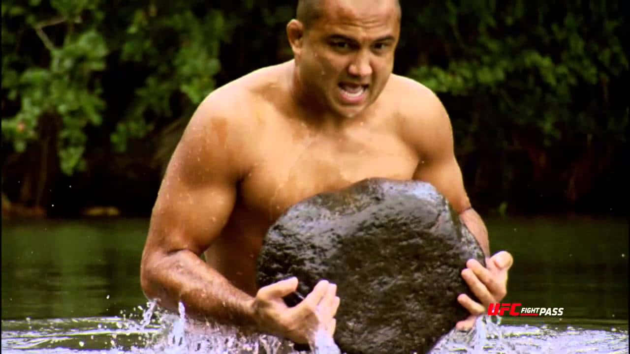 UFC 桌布 壁紙 — 一個肌肉發達、表情兇猛的男人從被綠色樹葉包圍的渾水中舉起一塊巨大的黑色岩石。