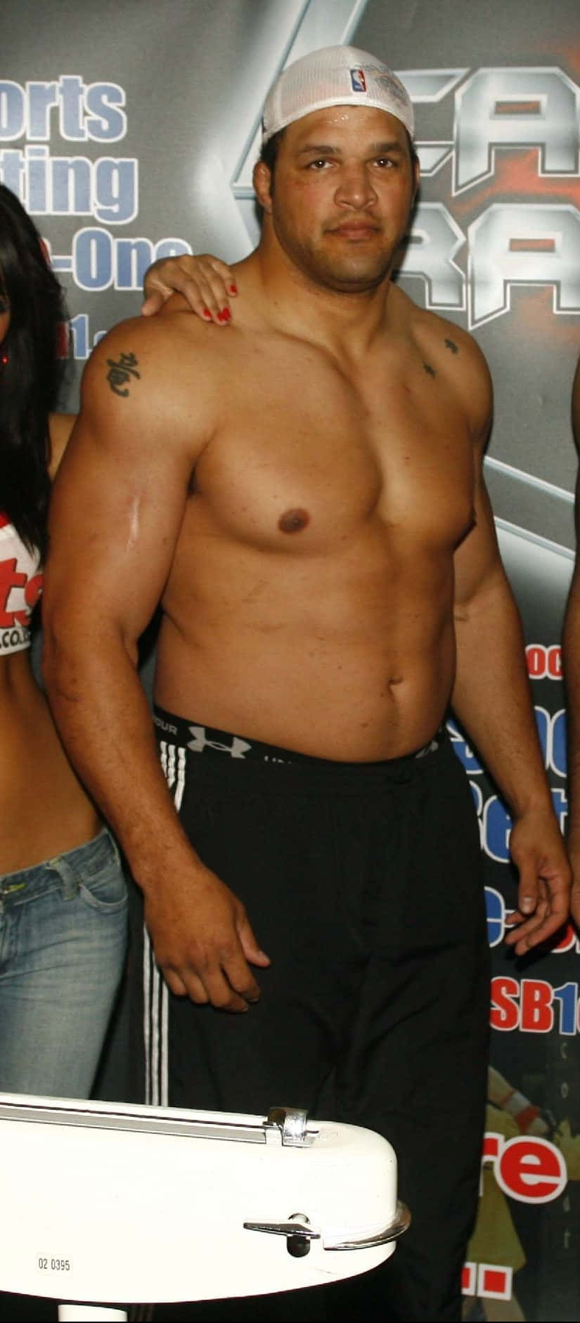 MMA 桌布 壁紙 — 一個肌肉發達、赤裸上身、手臂有刺青、戴白色帽子的男人，自信地站在體育賽事中，面前有一個磅秤。