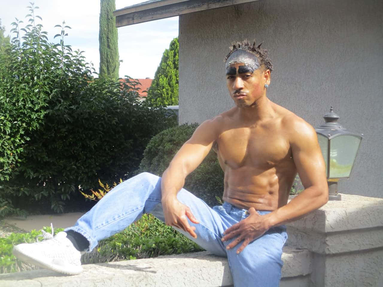 Ricardo Milos 桌布 壁紙 — 一個肌肉發達、赤裸上身、臉上塗有顏料的男子，在明亮的日光下隨意地坐在戶外的石牆上。