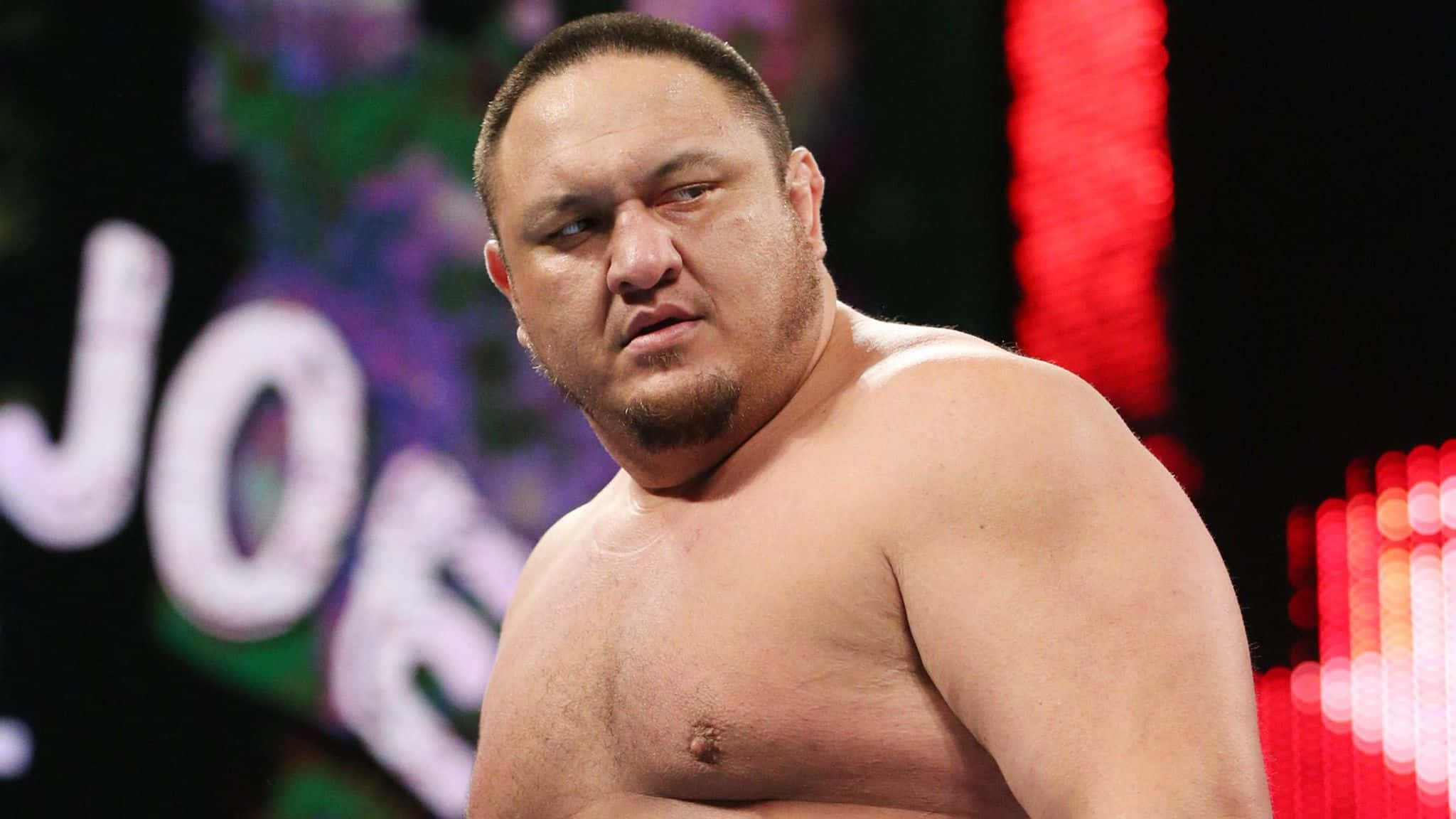 WWE 桌布 壁紙 — 一個肌肉發達、赤裸上身的摔角手，帶著專注的表情，站在一個燈火通明的競技場中，身後有色彩繽紛的模糊燈光。