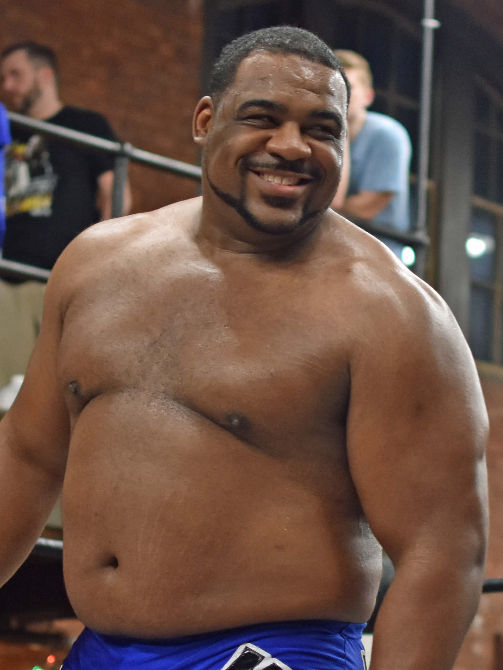 WWE 桌布 壁紙 — 一個肌肉發達、赤裸上身的男人，在明亮的燈光下自信地站在摔角擂台上，露出燦爛的笑容。