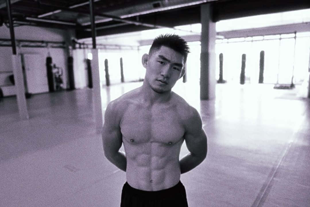 Athletic 桌布 壁紙 — 一個肌肉發達、赤裸上身的男人自信地站在一個簡陋的工業風健身房，有裸露的橫樑和明亮的窗戶。