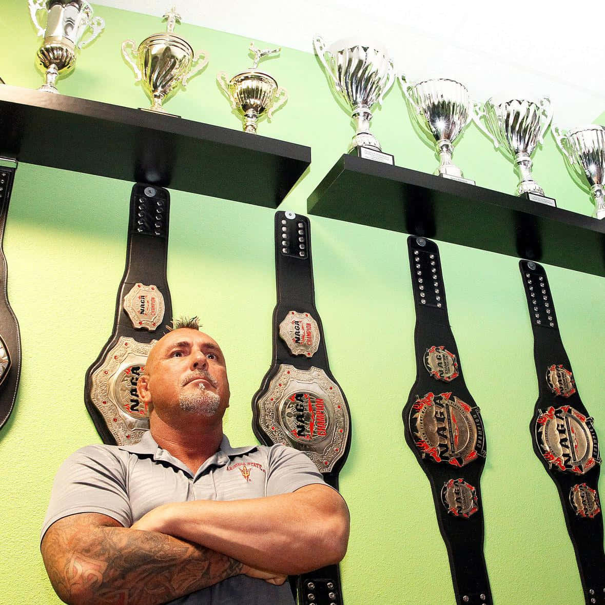 Trophy 桌布 壁紙 — 一個肌肉發達、身上有刺青的男人自豪地站在展示冠軍腰帶和閃亮獎盃的牆前。