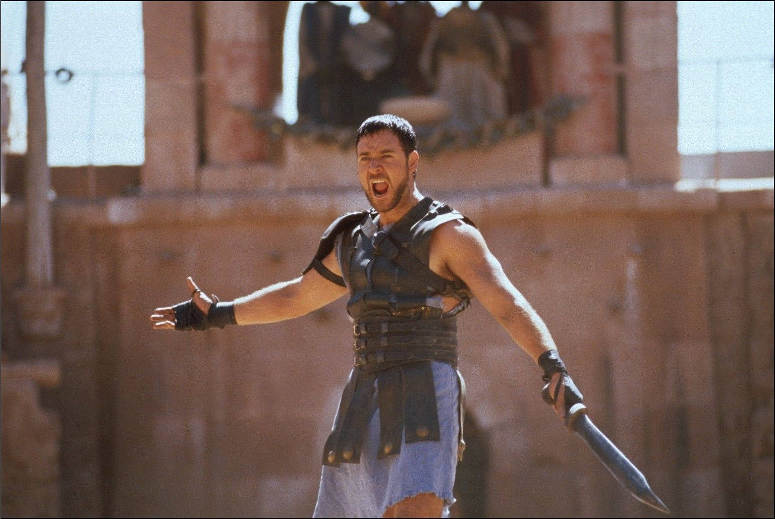 Gladiator 桌布 壁紙 — 一個肌肉發達、身穿盔甲的角鬥士，手持匕首，在陽光普照的古代競技場背景下發出勝利的咆哮。