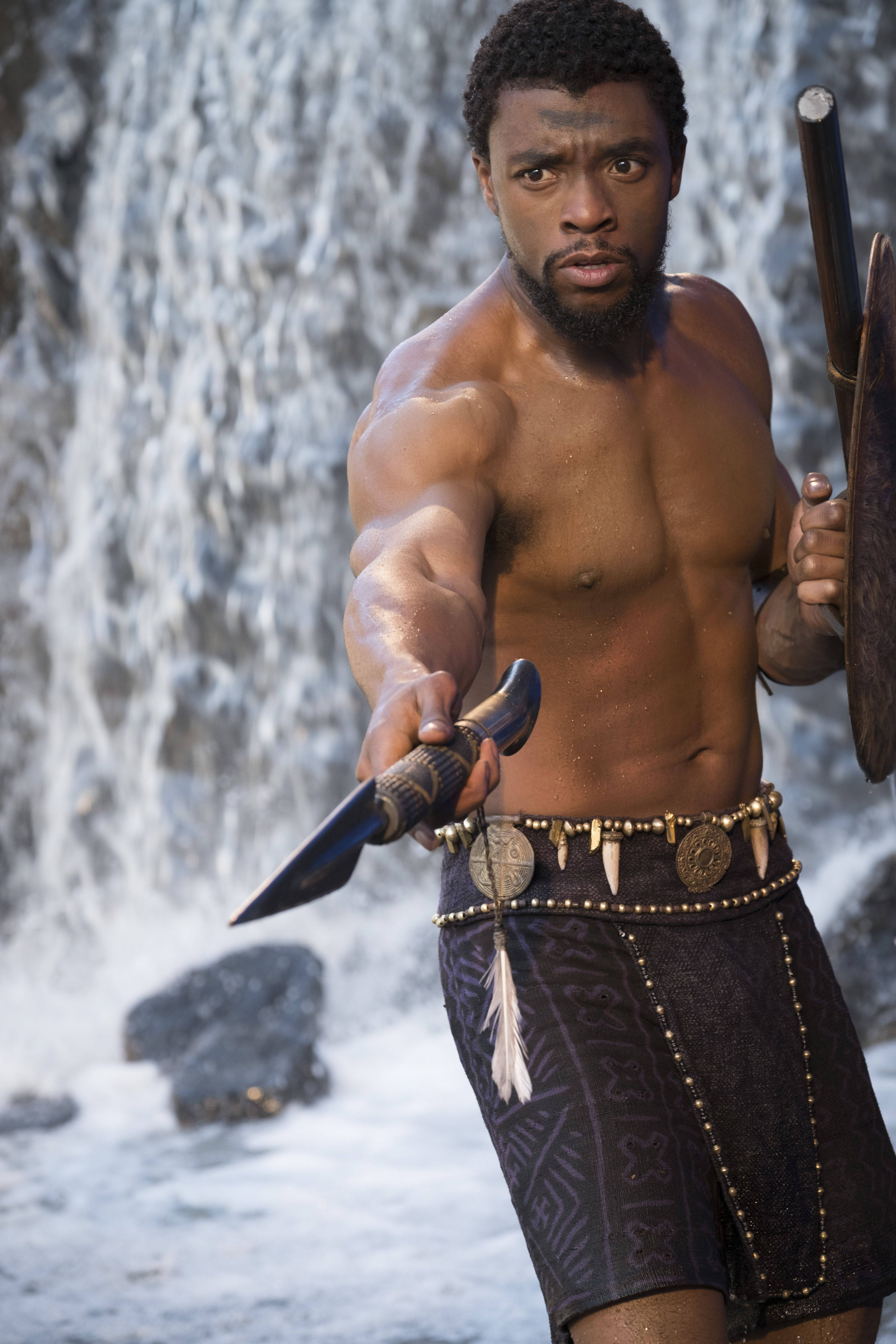 Chadwick 桌布 壁紙 — 一個肌肉發達、身穿部落服飾的男人，手持匕首和盾牌，挑釁地站在瀑布前。