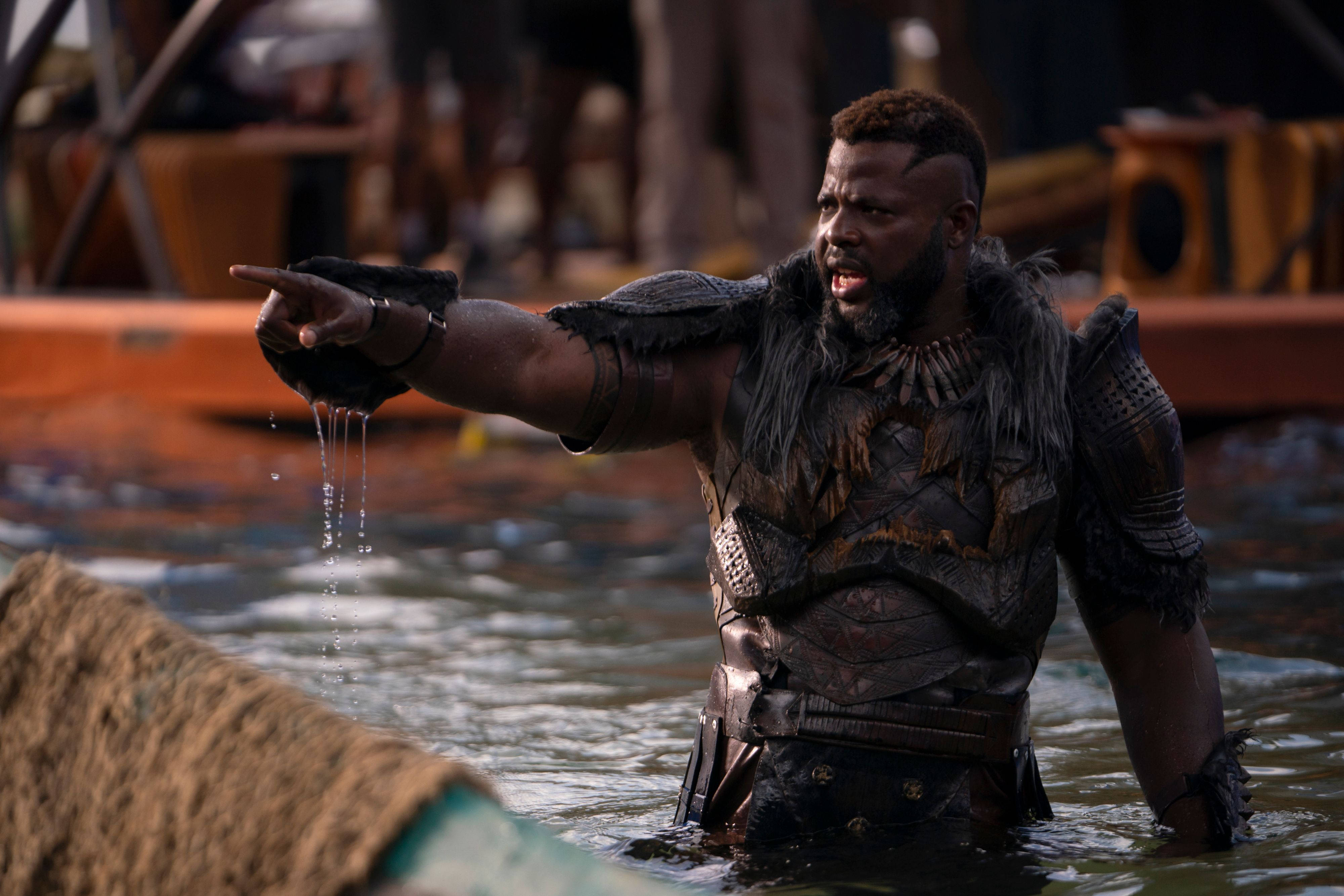 Wakanda 桌布 壁紙 — 一個肌肉發達、身穿部落盔甲的男人站在齊腰深的水中，手指向前方，水滴從他的手上滴落。