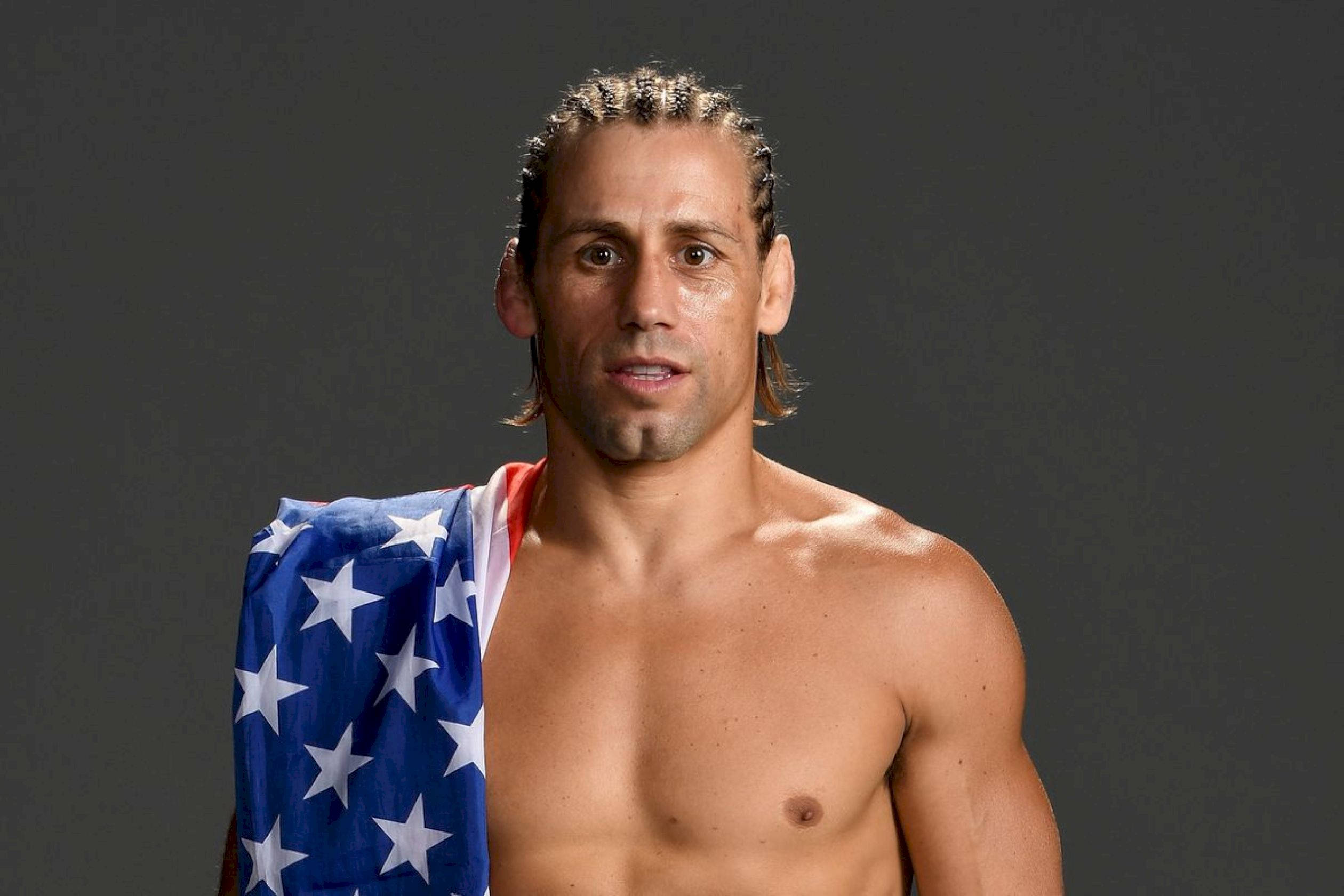 UFC 桌布 壁紙 — 一個肌肉發達、辮子頭髮的男人赤膊站在深灰色背景前，肩上披著美國國旗。