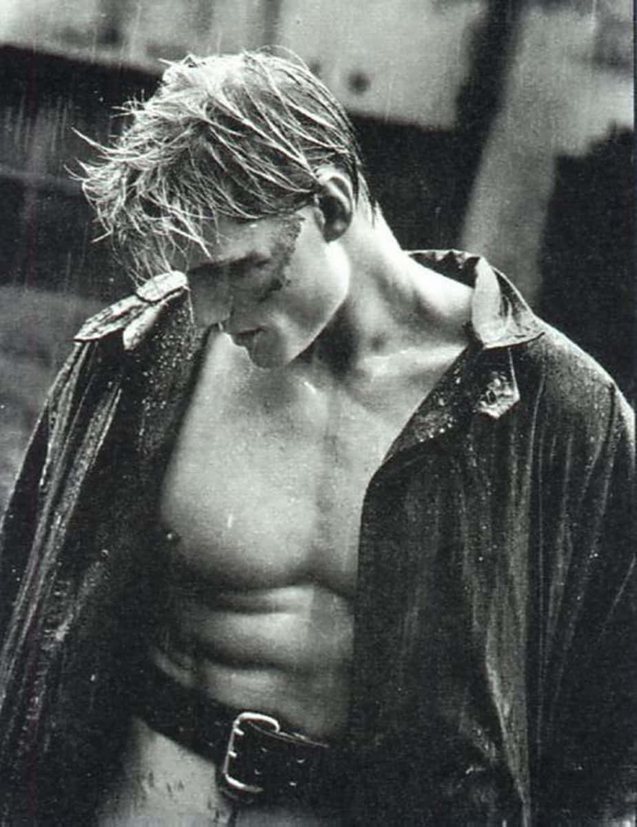 Dolph Lundgren 桌布 壁紙 — 一個肌肉發達、頭髮濕透的男人赤裸上身站在雨中，敞開的夾克緊貼著他的軀幹，水珠沿著他的臉和胸膛滴落。