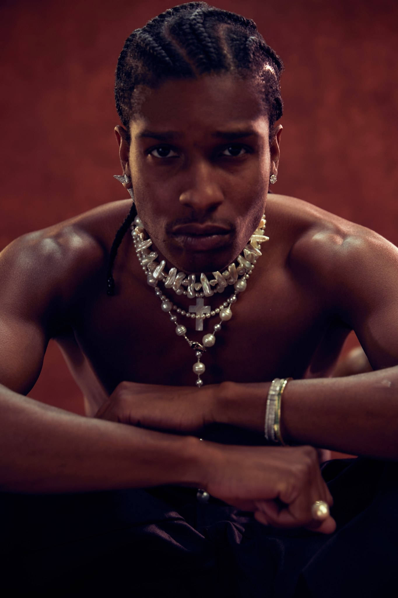 rocky 桌布 壁紙 — 一個肌肉發達、頭髮編成辮子的男人，在深紅色背景下佩戴著多層珍珠和十字架項鍊，眼神銳利。