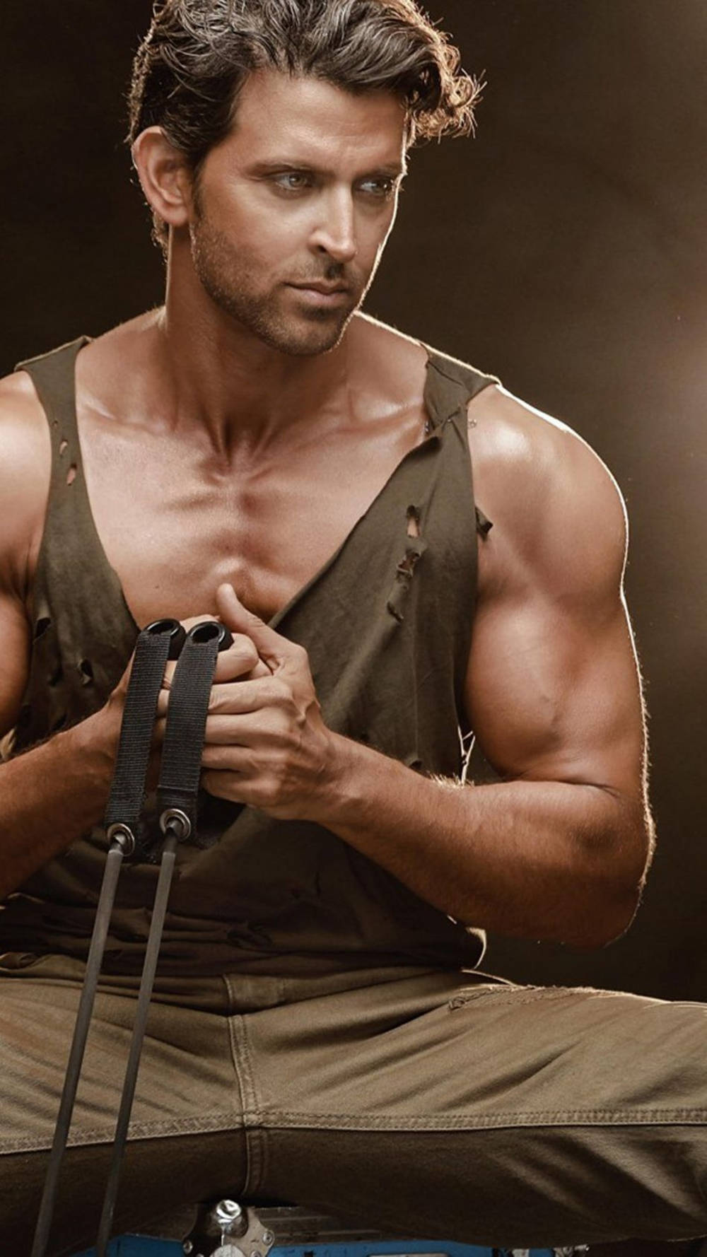 Workout 桌布 壁紙 — 一個肌肉發達、黑髮帶鬍渣的男人穿著破爛的橄欖綠背心，在昏暗的攝影棚中抓著阻力帶。