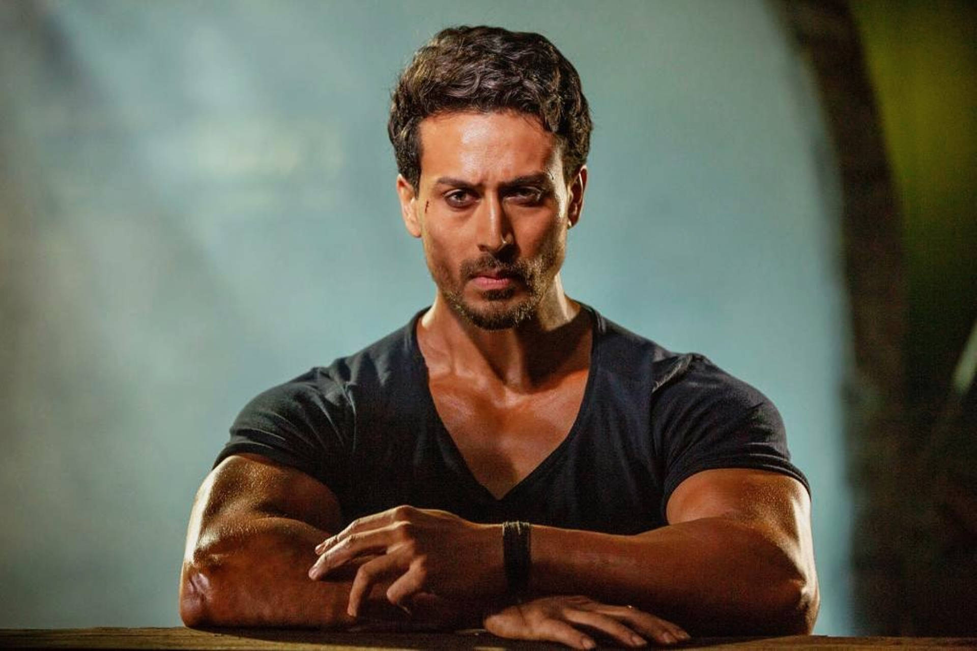 Tiger Shroff 桌布 壁紙 — 一個肌肉發達、黑髮留鬍的男人在昏暗的環境中，將手臂擱在木質表面上，眼神銳利地凝視。