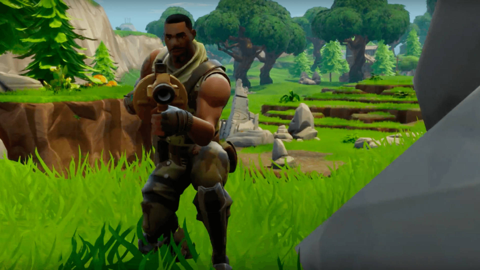 電玩遊戲 桌布 壁紙 — 一個肌肉發達的Fortnite角色蹲在充滿活力的綠色草地上，在岩石地形和樹木附近手持武器。